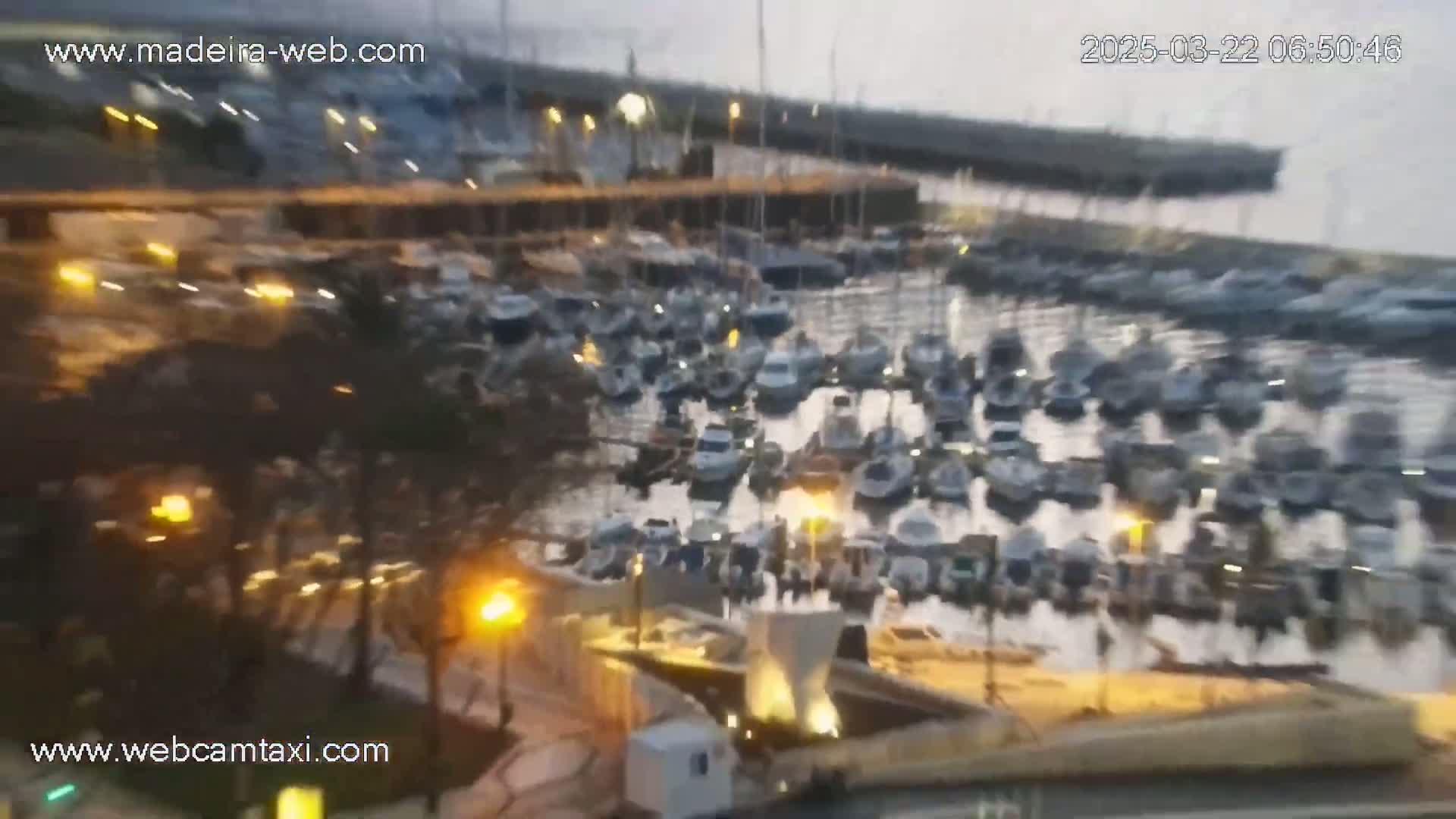 A harbor filled with many boats is seen at twilight, with some lights visible on land and possibly a slight haze in the air.