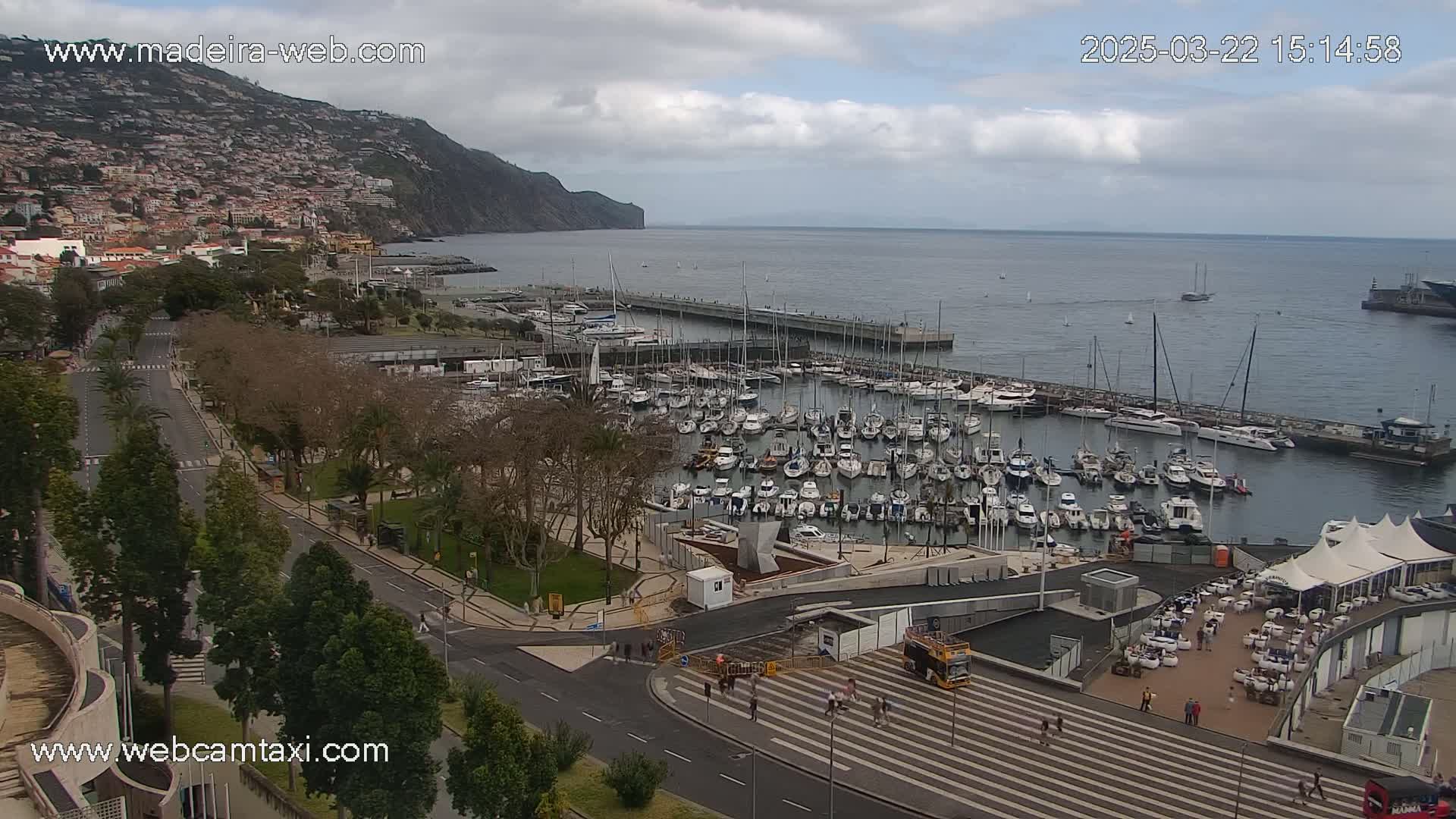 A marina filled with numerous boats is situated beside a city nestled against a steep, dark-green mountain, all under a partly cloudy sky.
