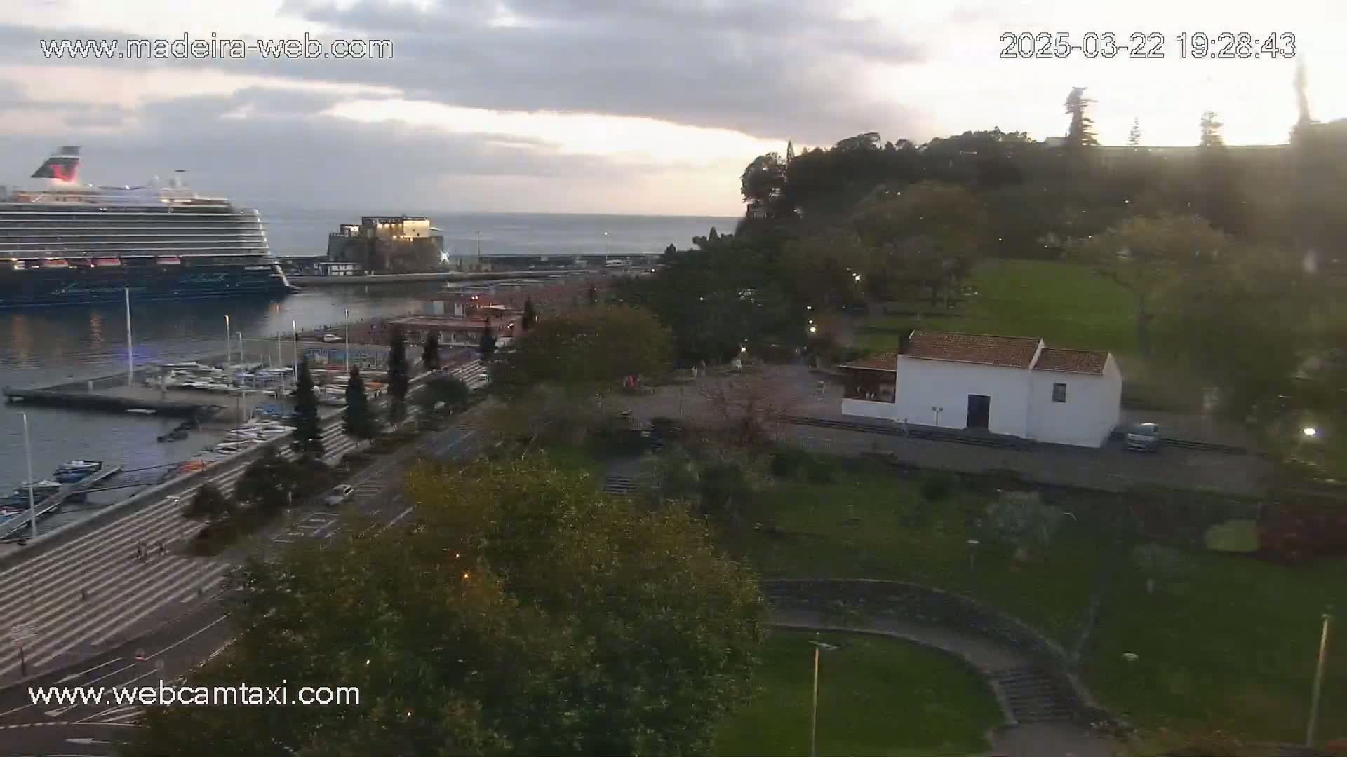 A large cruise ship is docked at a harbor next to a park with a small white building, under a cloudy evening sky.