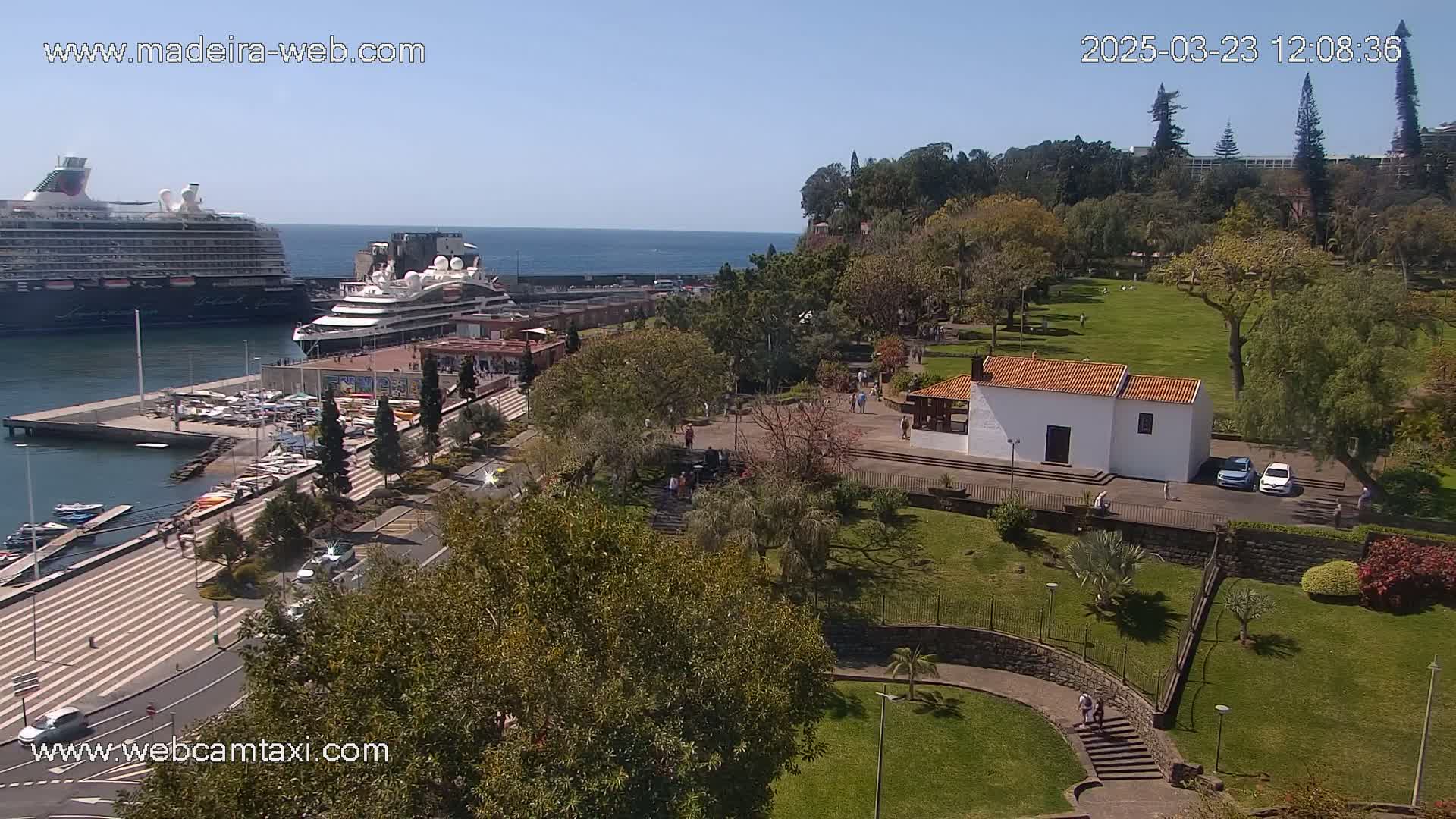 An elevated view of a harbor with two cruise ships, a paved waterfront area with cars, and a park with a small white building on a sunny day.