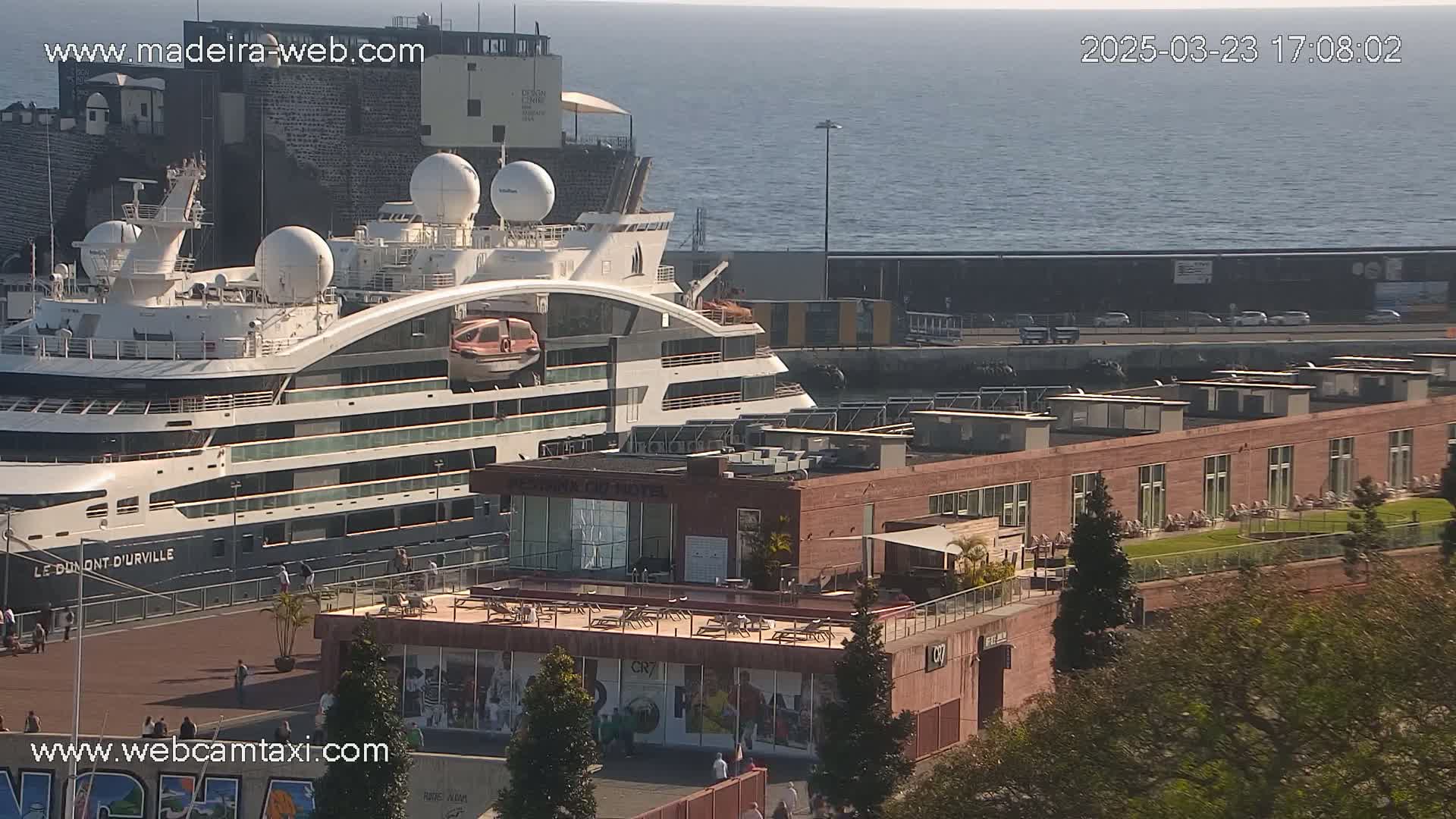 A large cruise ship is docked at a port on a sunny day, next to a reddish-brown building with a rooftop patio.