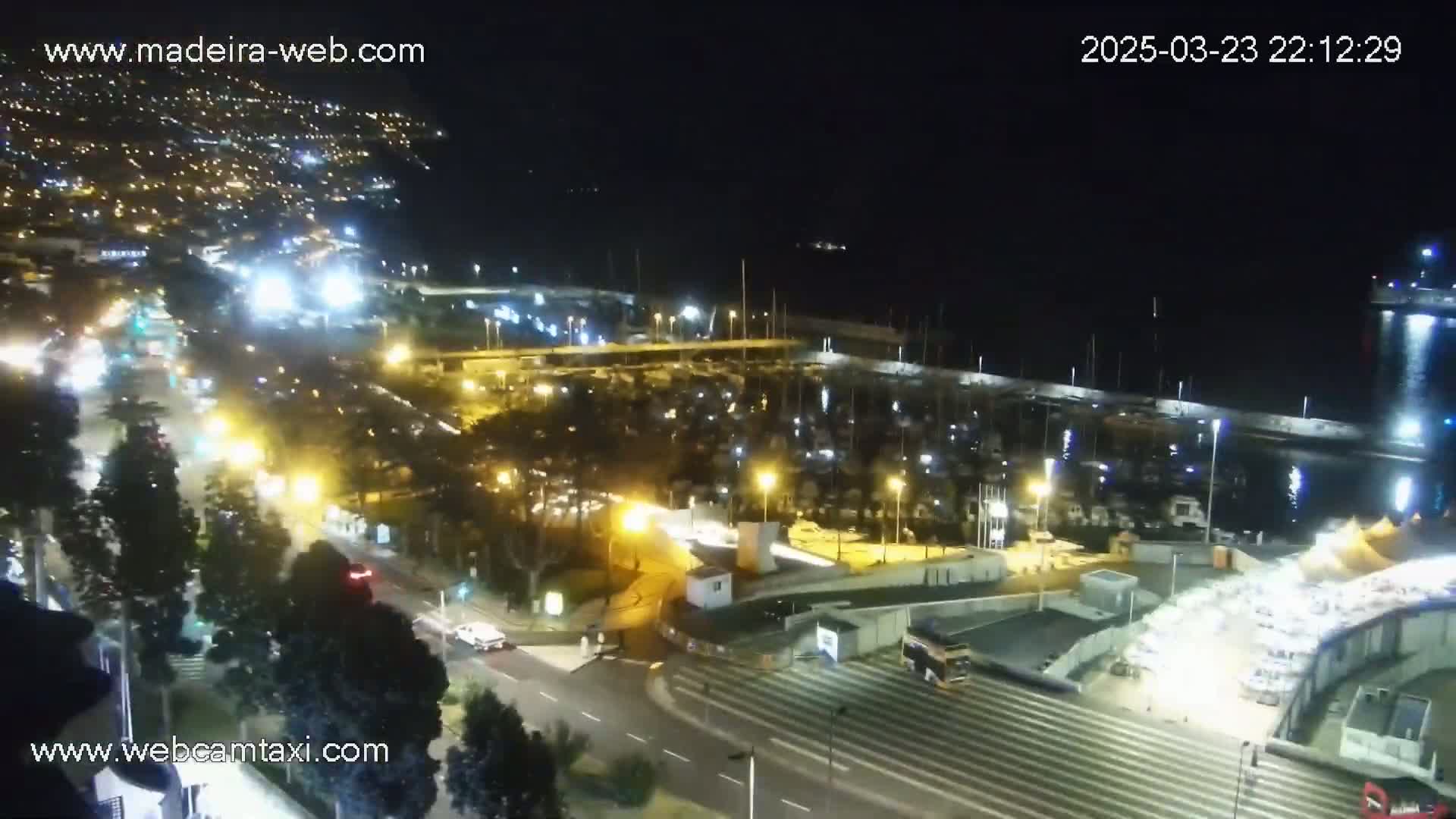 A nighttime, aerial view shows a marina filled with boats, a road with moving vehicles, and a city illuminated with lights in the background.