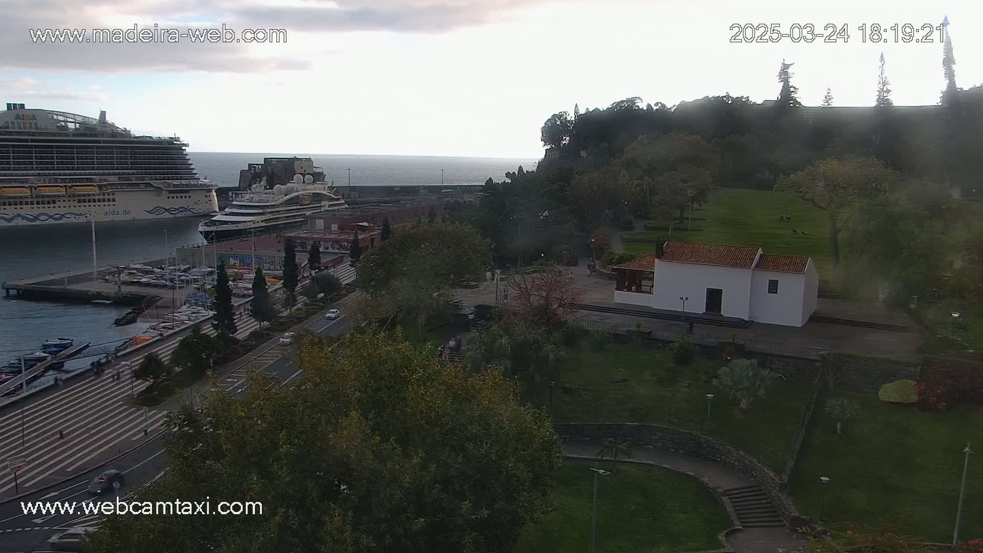 An overcast day shows a cruise ship docked at a port, with a view overlooking a park containing a small white building and a road leading down to the water.