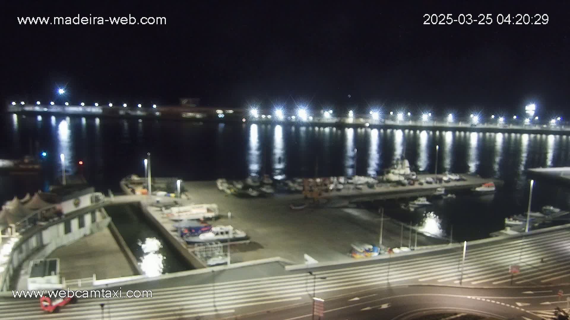 A nighttime view of a harbor filled with boats, illuminated by bright lights reflecting on the calm water.