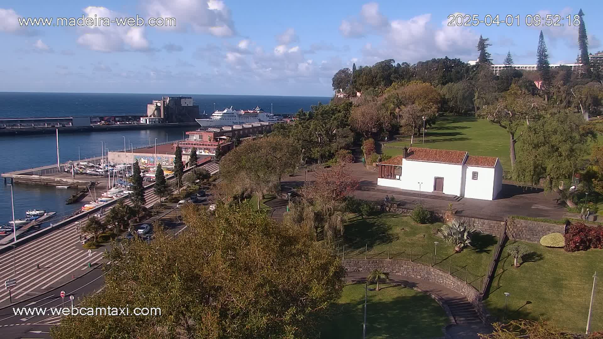 A harbor with a cruise ship, marina, and waterfront promenade is adjacent to a park with a small white building under a partly cloudy sky.