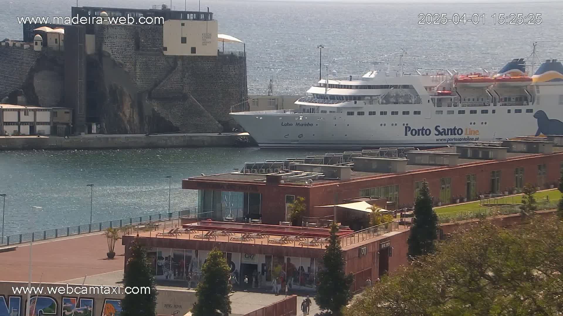 A large passenger ferry is docked at a harbor next to a stone fortress, on a partly sunny day with calm water.