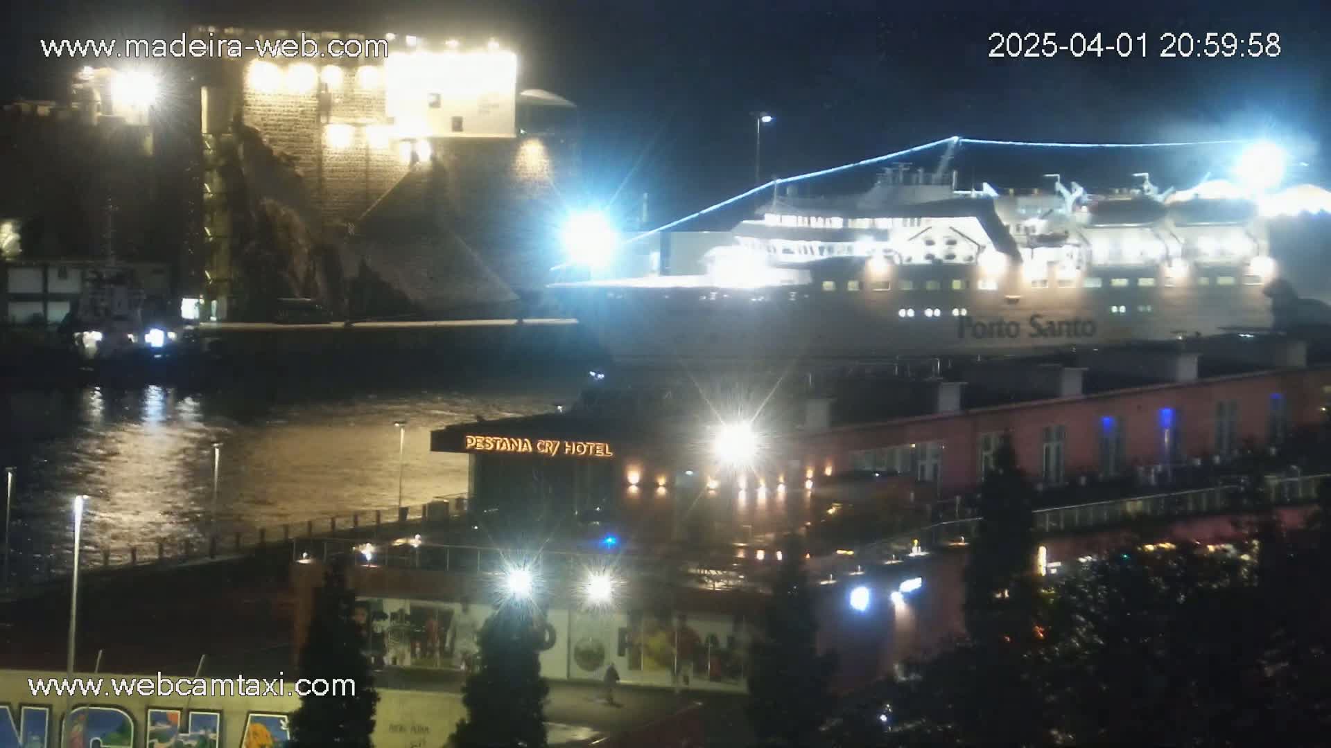 A large ferry is docked at a harbor at night, next to a stone structure and a hotel, under slightly overcast conditions.