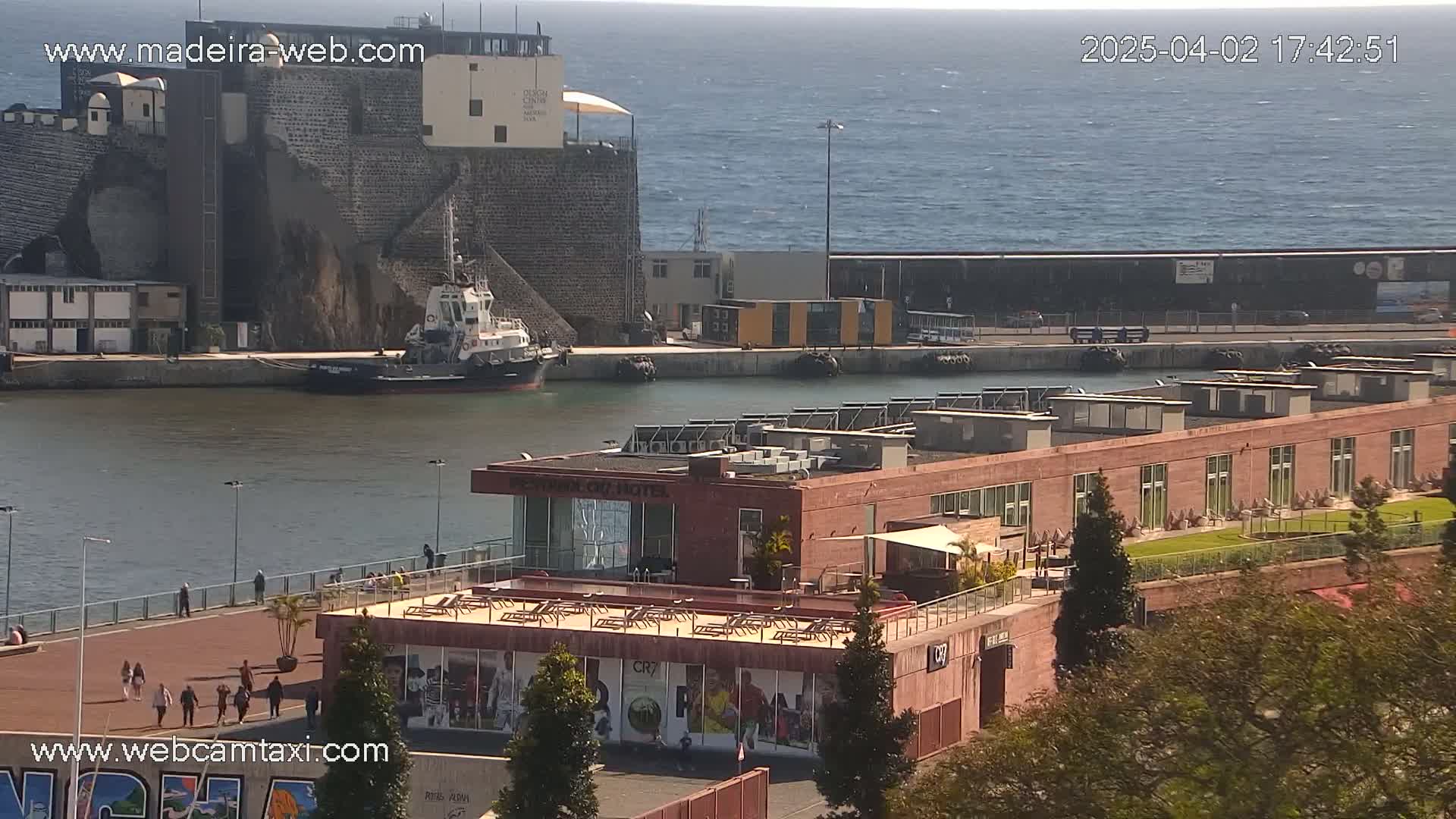 A harbor scene on a sunny day shows a tugboat near a stone structure, a long building with a rooftop patio, and calm water leading to a blue ocean.