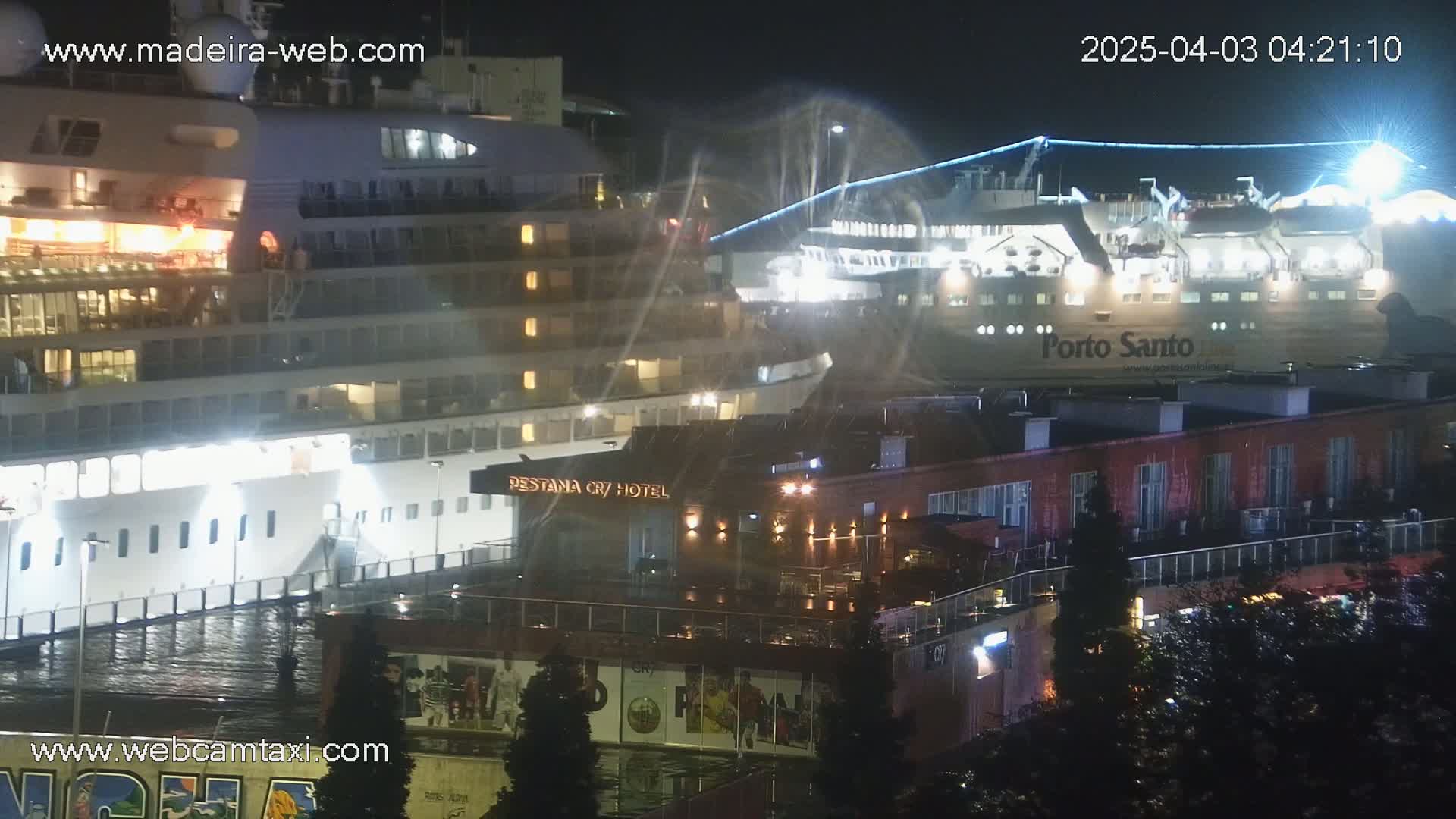 At night, a large cruise ship and a ferry are docked near a hotel and other buildings, with light rain or spray visible.