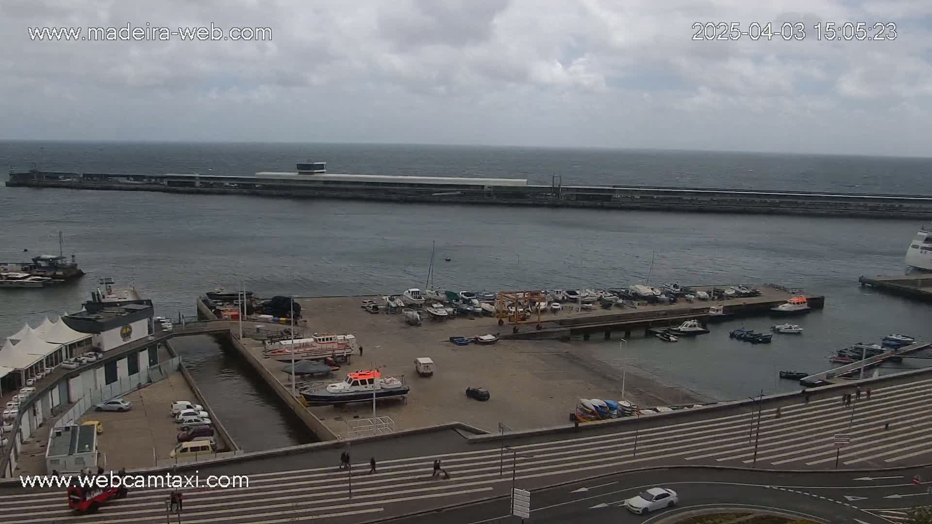 An overcast day shows a harbor filled with numerous small boats and a few larger vessels near a waterfront building and a long pier stretching out to the ocean.