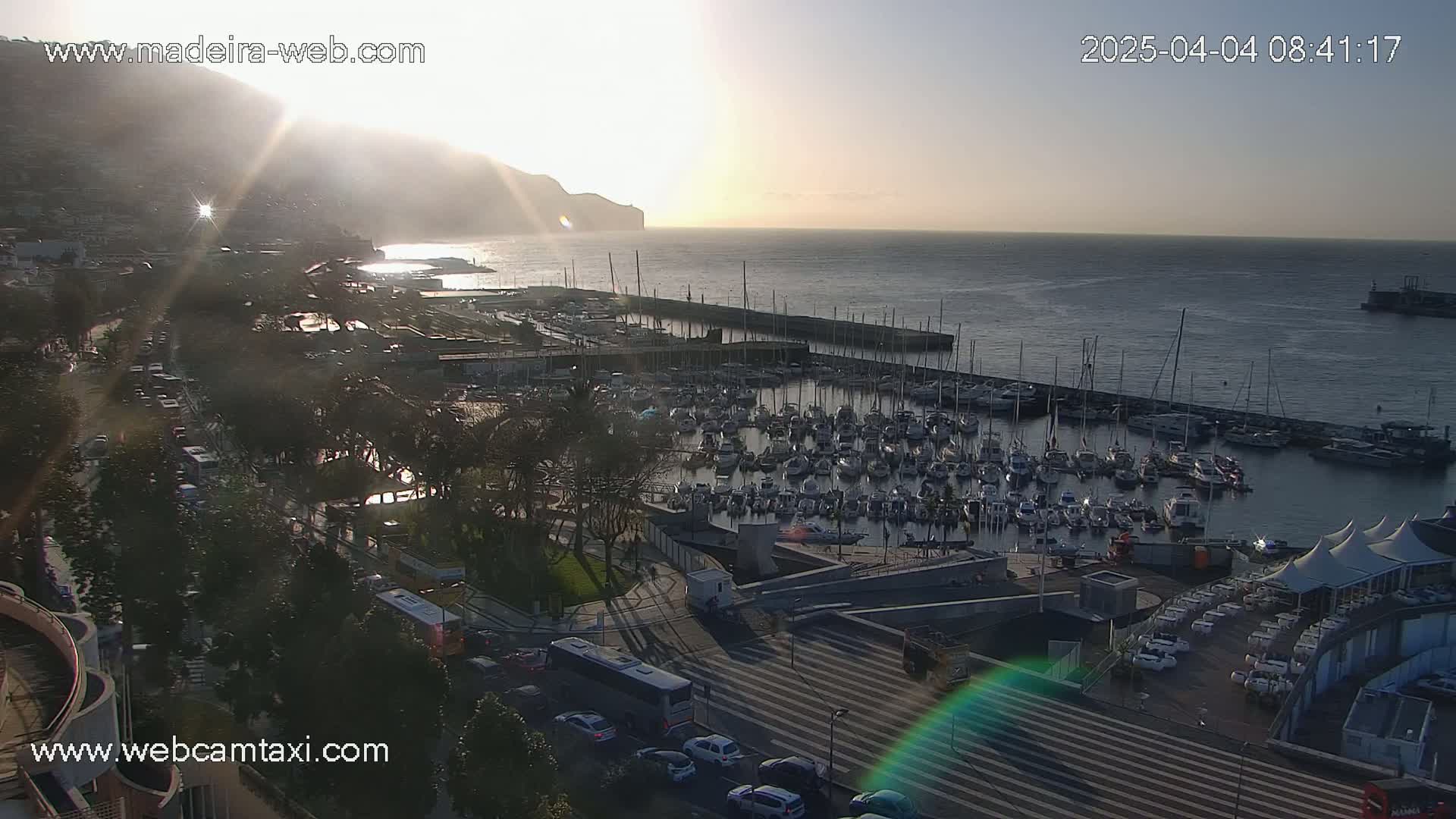 A sunlit marina filled with numerous boats is situated near a coastal town at sunrise, with a mountain visible in the background.