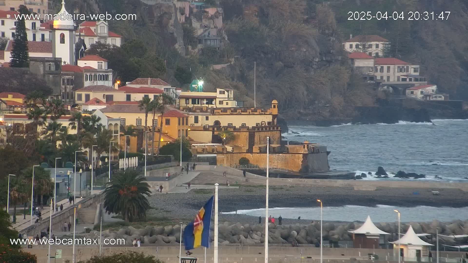 A coastal town at dusk, with a stone fortress overlooking a dark-sand beach and a somewhat choppy ocean.