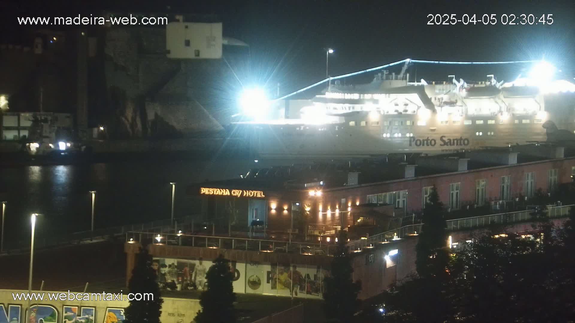 A large, brightly lit ferry is docked at a harbor at night next to a hotel and other buildings.