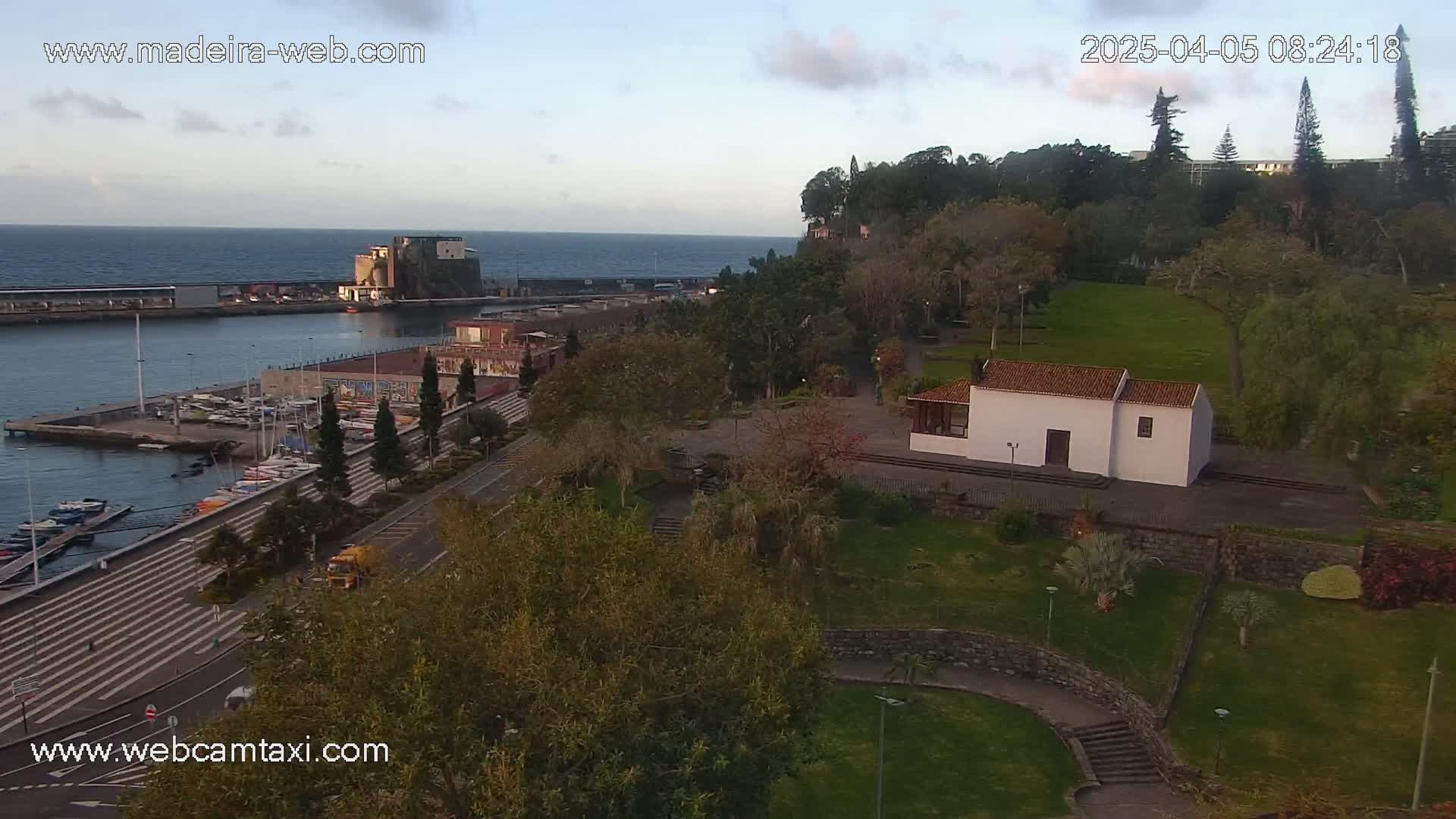 An elevated view of a harbor with boats, a waterfront promenade, a park with a small white building, and a hillside with trees, under a partly cloudy dawn sky.
