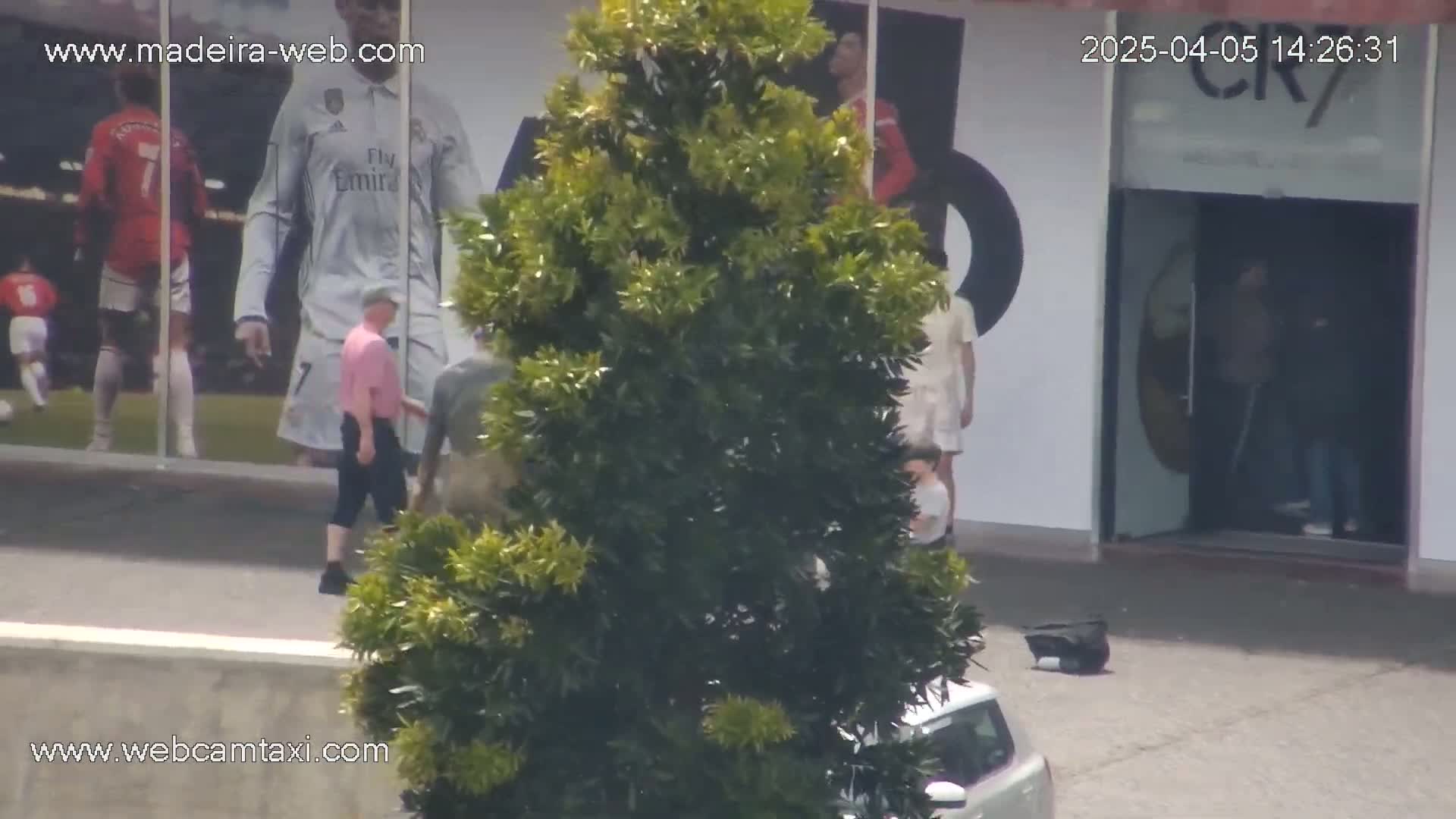 A large green bush partially obscures a view of several people walking near a building with large images of soccer players on its exterior, on a sunny day.