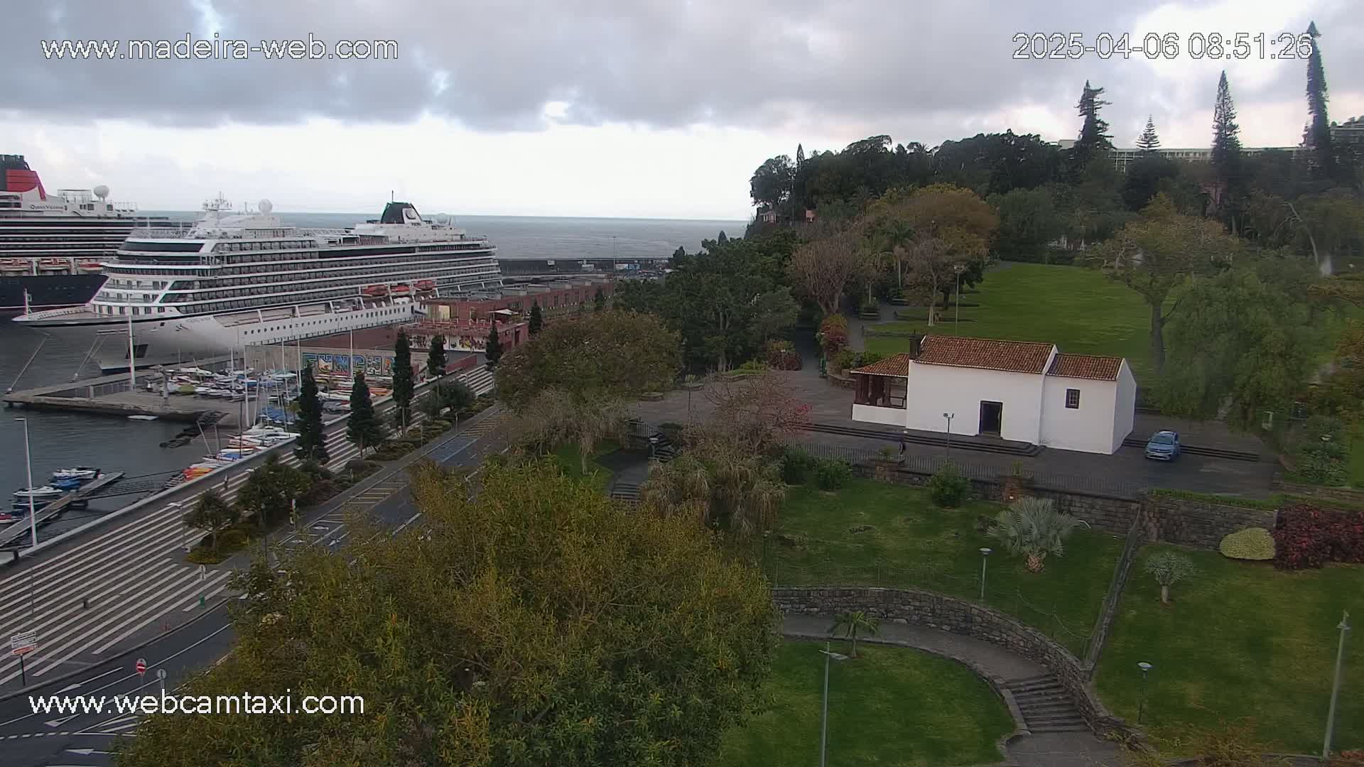 A large cruise ship is docked at a harbor next to a park with a small white building, under a cloudy sky.