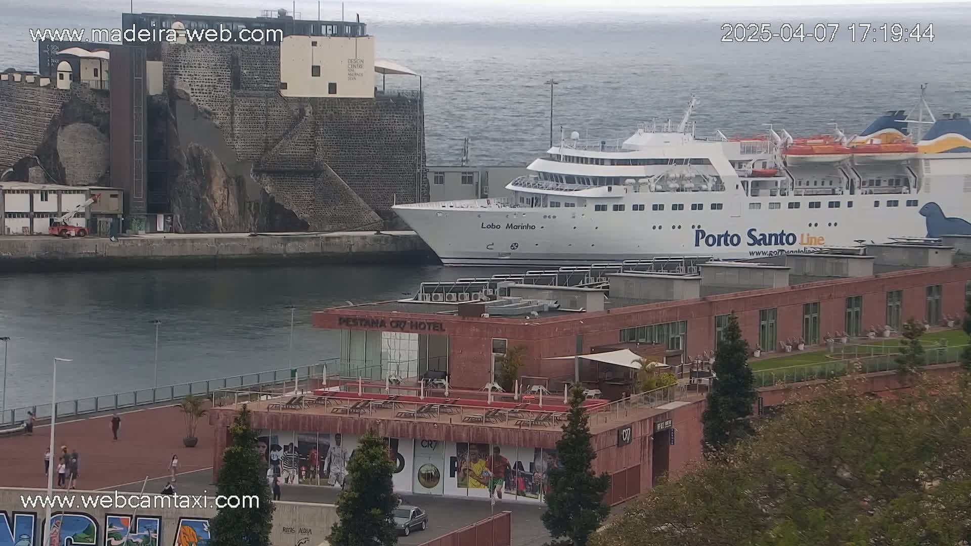A large white ferry is docked at a harbor next to a building with a rooftop deck, under a mostly cloudy sky.