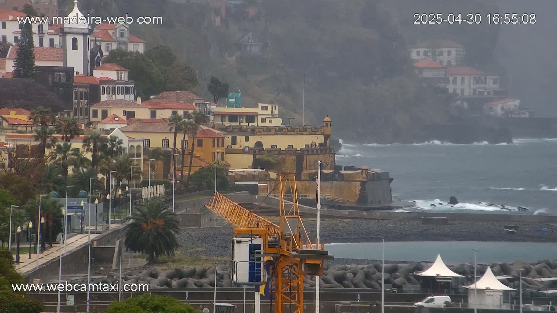 A coastal town with colorful buildings, a stone fort, and a construction crane is shown on an overcast day with a choppy ocean.