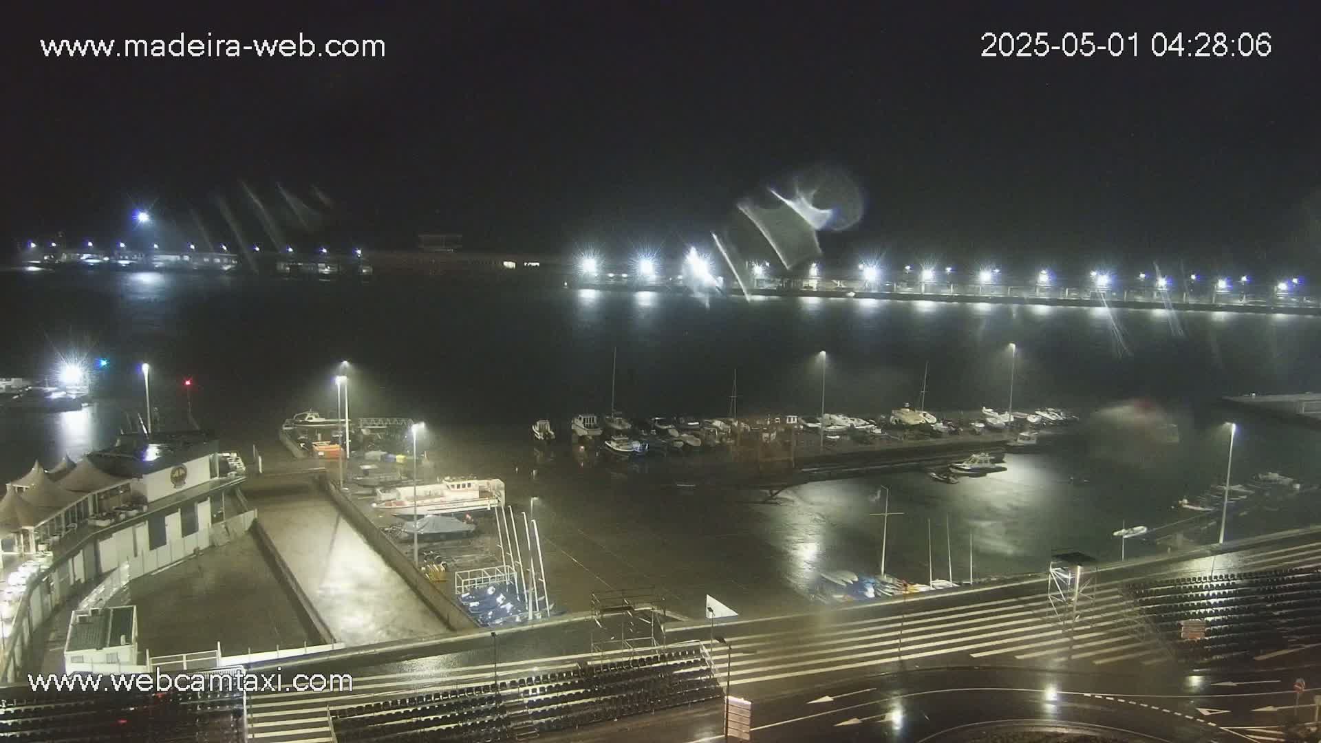 A nighttime view of a marina with many boats docked, under rainy conditions, showing a building and empty seating areas nearby.