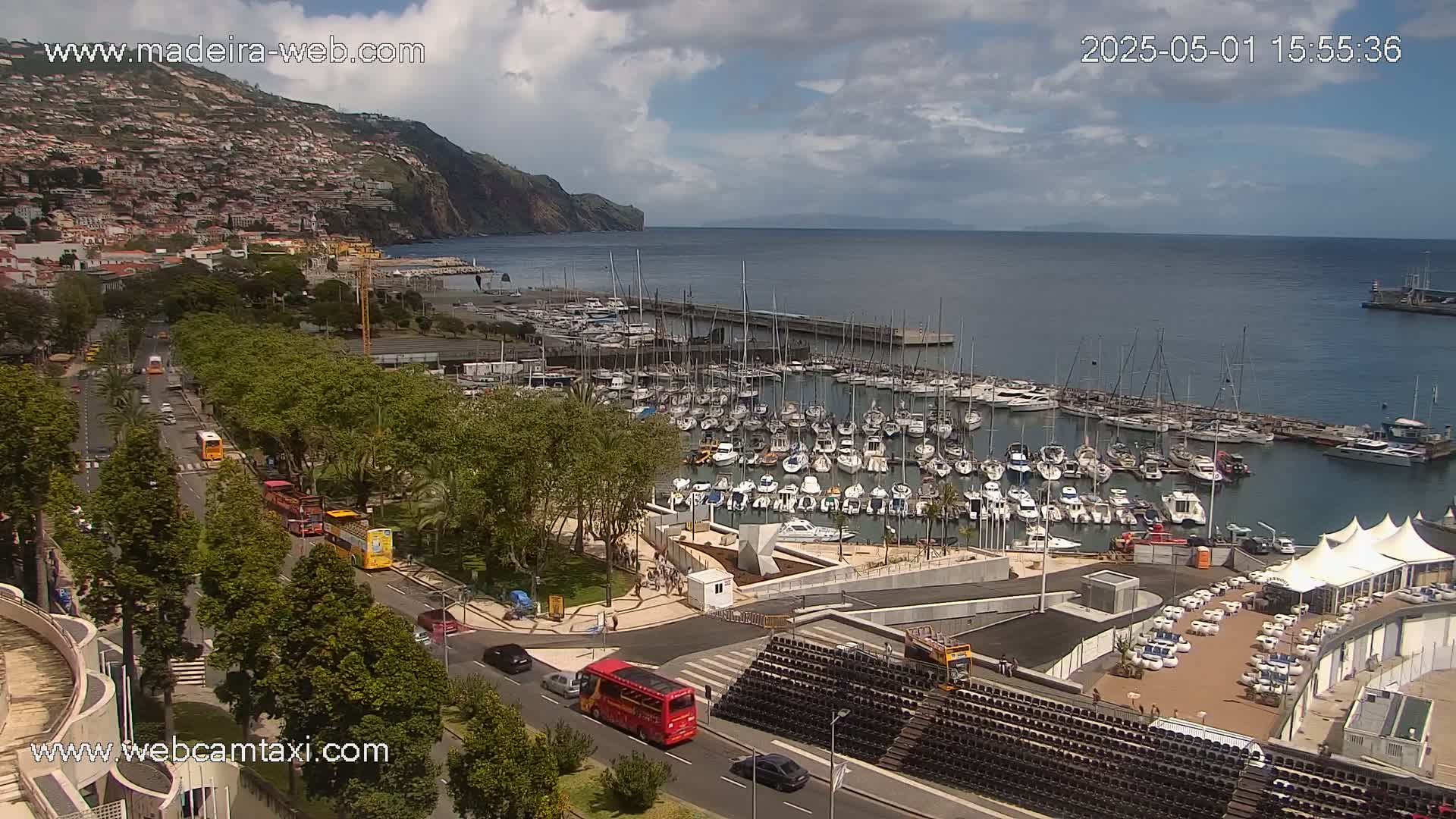 A harbor filled with many sailboats and motorboats is situated below a hillside town, under a partly cloudy sky.