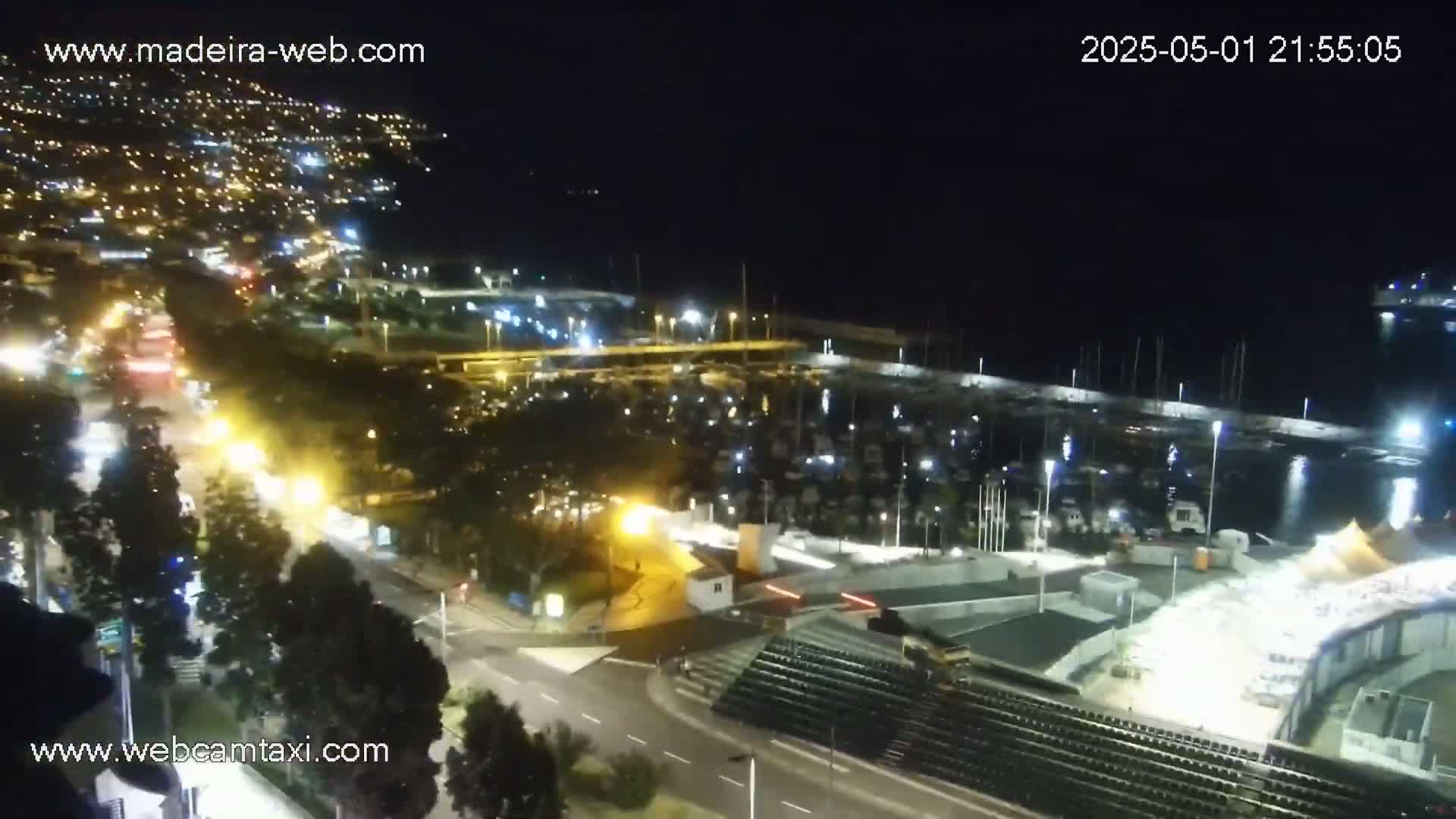 A nighttime, aerial view of a marina filled with boats, adjacent to a roadway with traffic, and a hill with illuminated buildings in the background.