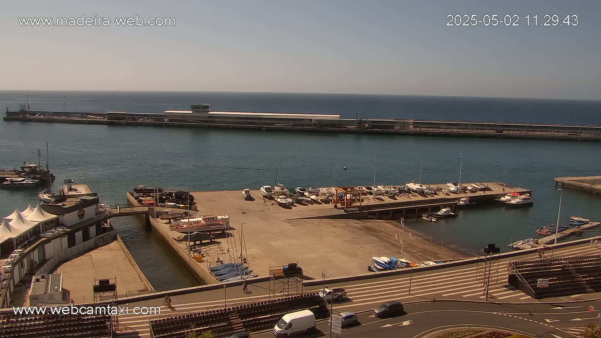 A harbor with numerous boats docked, a long pier extending into a calm ocean under a clear, sunny sky.