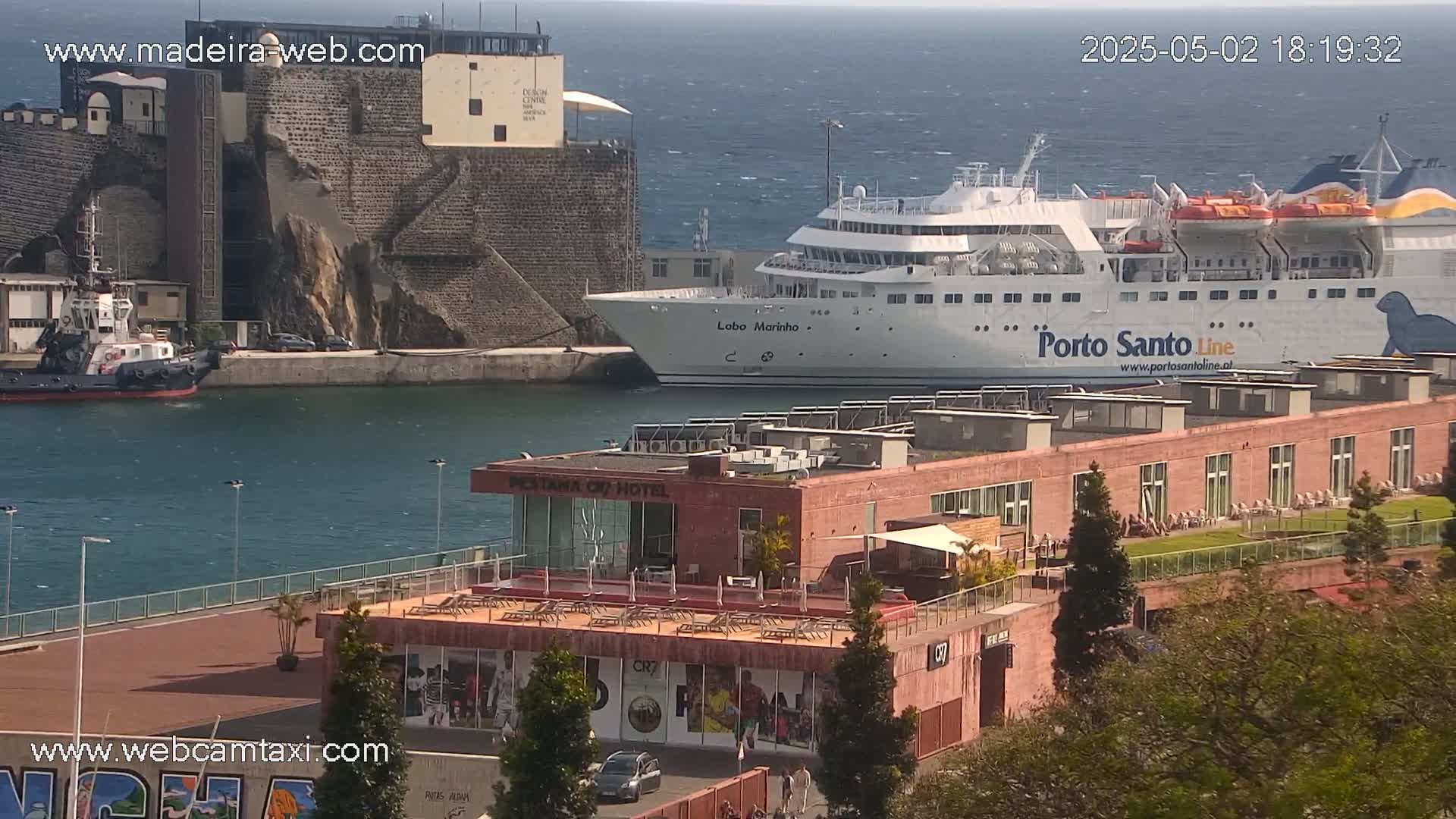 A large ferry is docked at a harbor next to a stone fortress, with a smaller tugboat nearby, under a mostly sunny sky with a light breeze visible on the water.