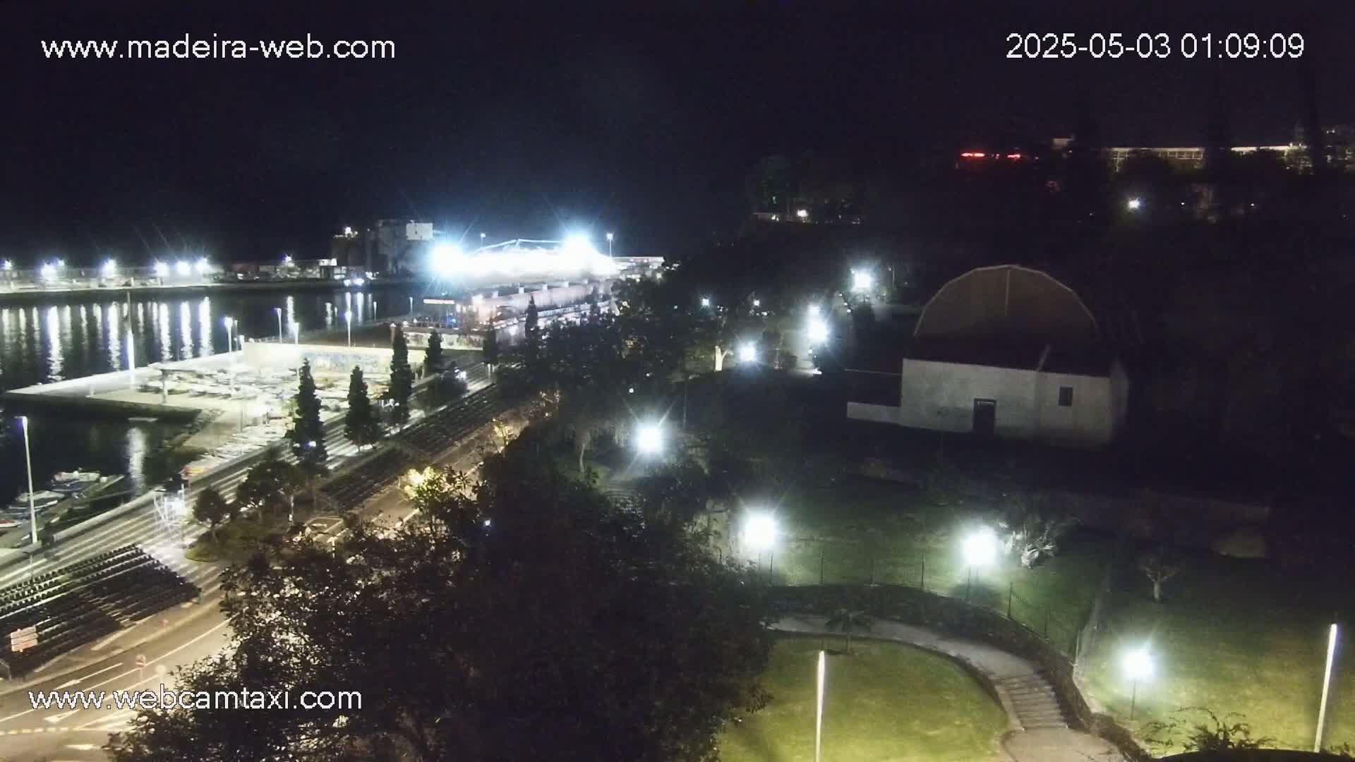 A nighttime view of a harbor with boats, a stadium, a building with a domed roof, and a park illuminated by lights.