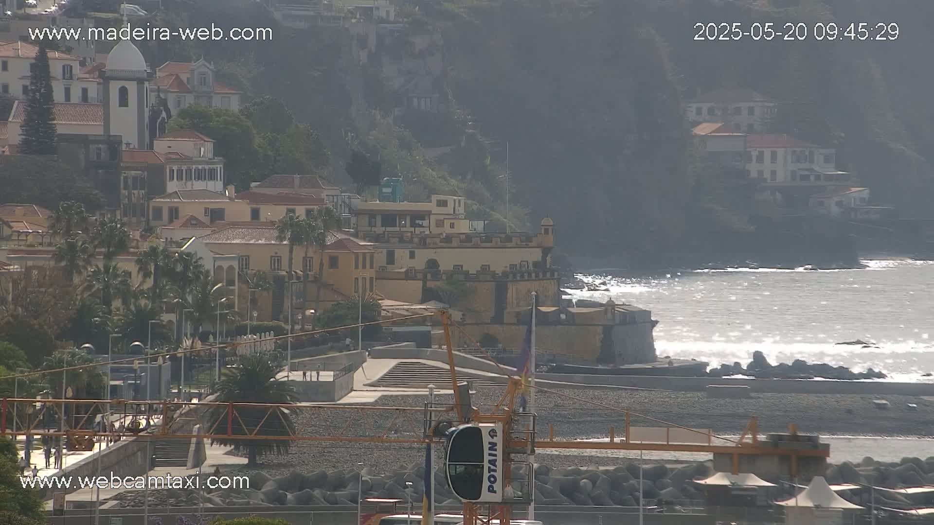 A coastal town with a mix of buildings, including a church, overlooks a rocky shore with a slightly wavy ocean under a sunny sky.