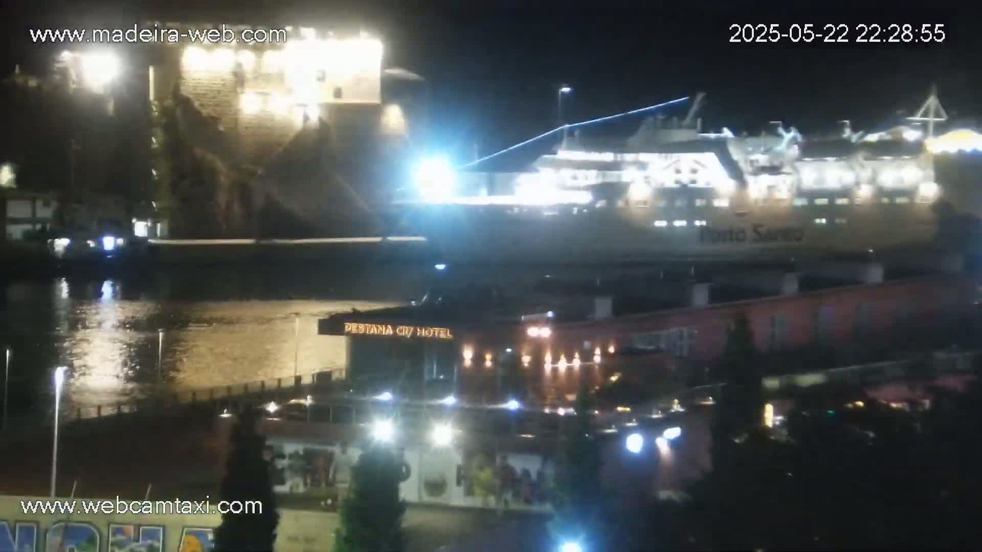A large ferry is docked at night next to a stone building and hotel, illuminated by lights reflecting on the calm water.