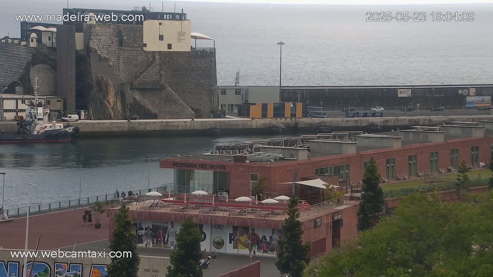 A harbor scene on an overcast day shows a tugboat in the water near a brick hotel with a rooftop patio, adjacent to a stone structure and a roadway.