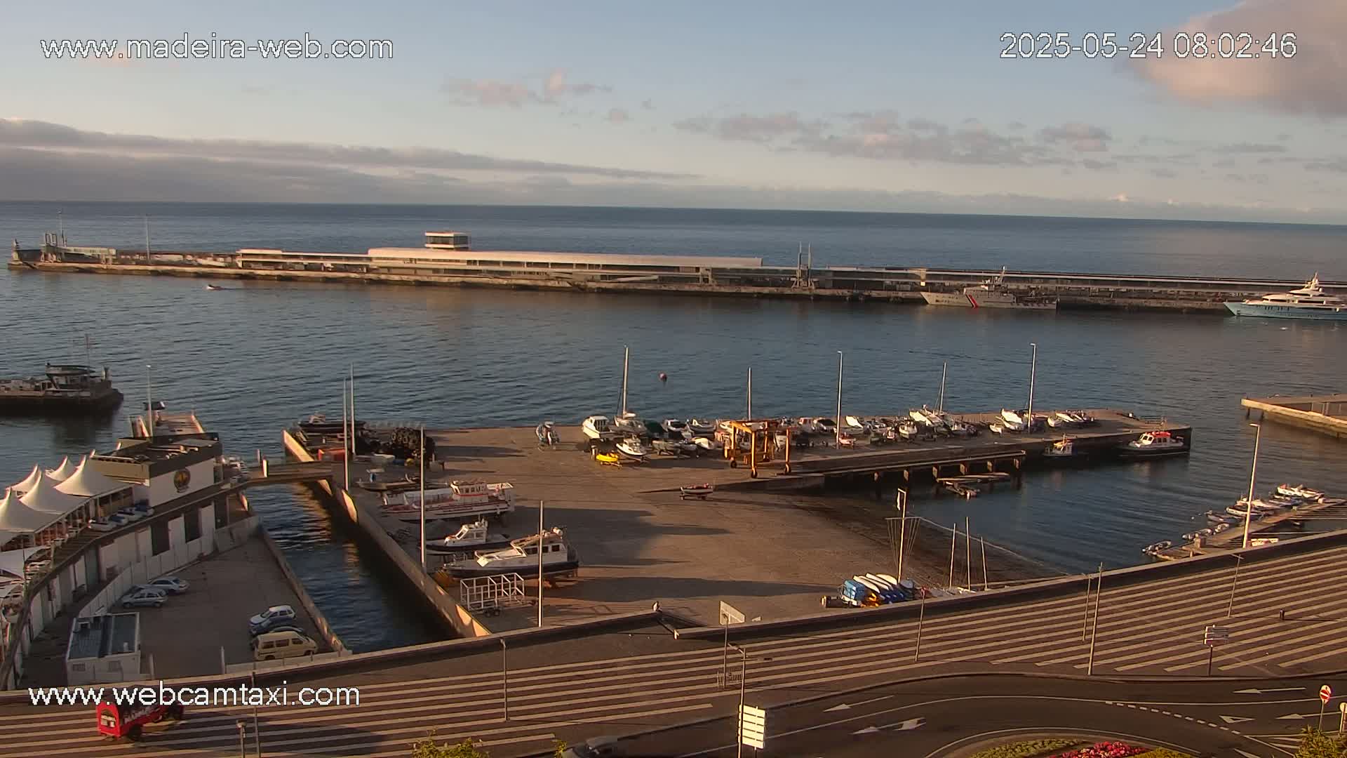 An aerial view of a harbor with numerous boats docked, a long pier extending into the calm water under a partly cloudy morning sky.