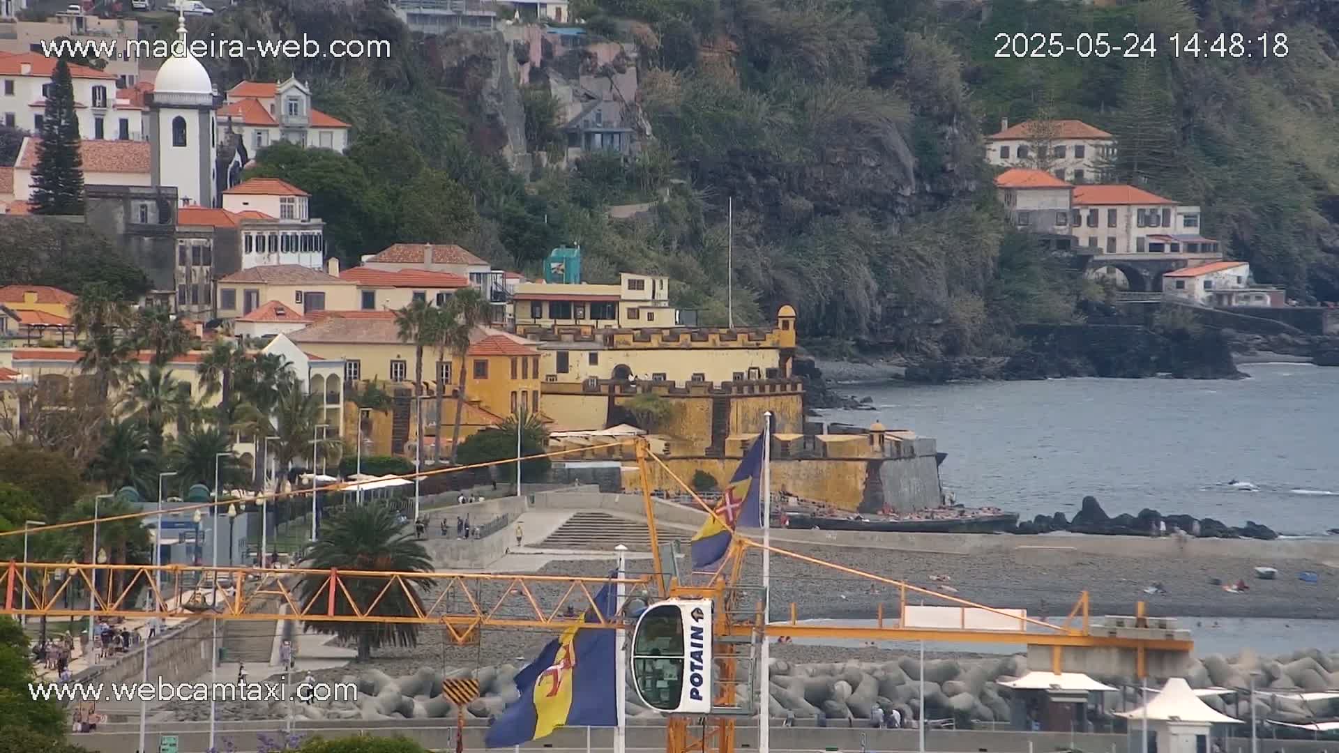 A coastal town with colorful buildings, a stone fortress, and a pebble beach, under partly cloudy skies.