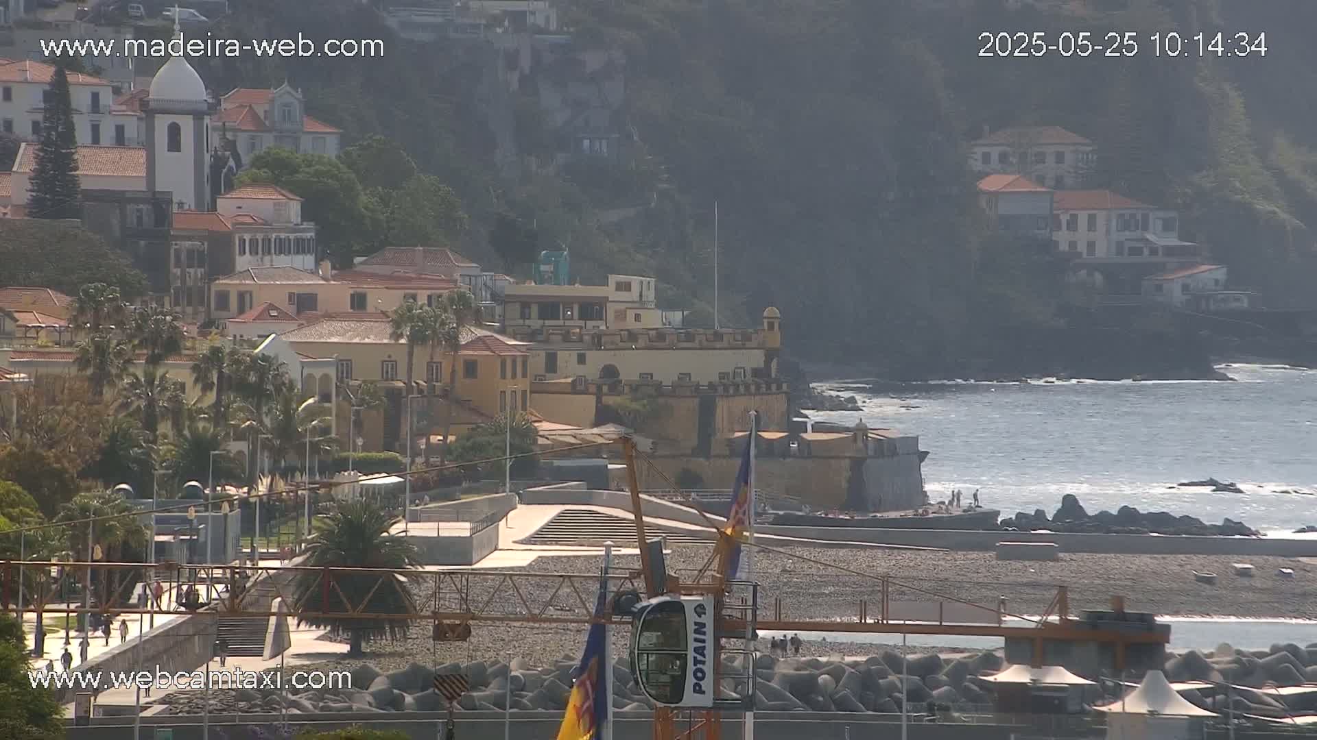 A coastal town with a mix of old and new buildings sits on a hillside overlooking a rocky beach and calm ocean under a mostly sunny sky.