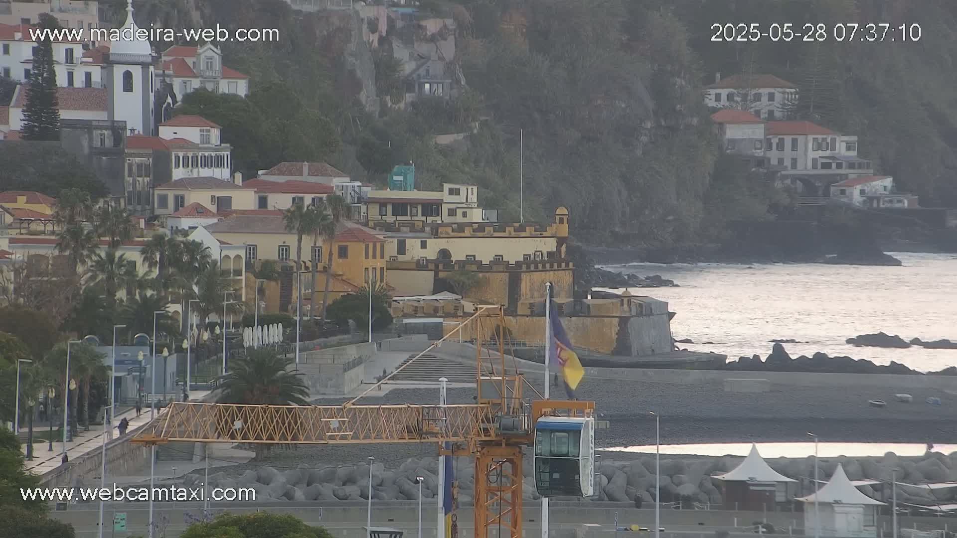 A coastal town with a stone fort and a construction crane is visible under overcast skies.