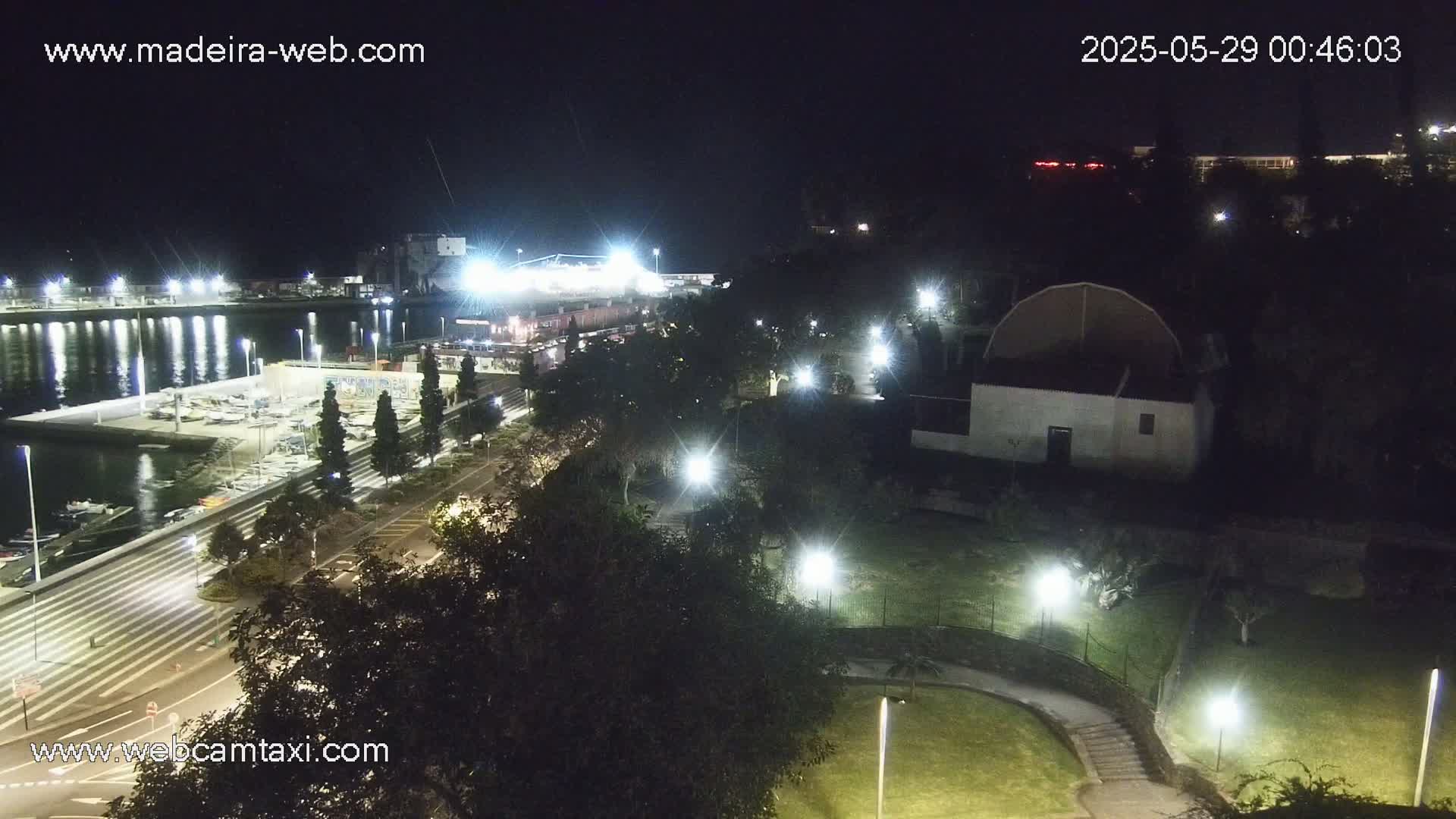 A nighttime aerial view of a marina, a roadway, and a park with several buildings, all illuminated by artificial lights.