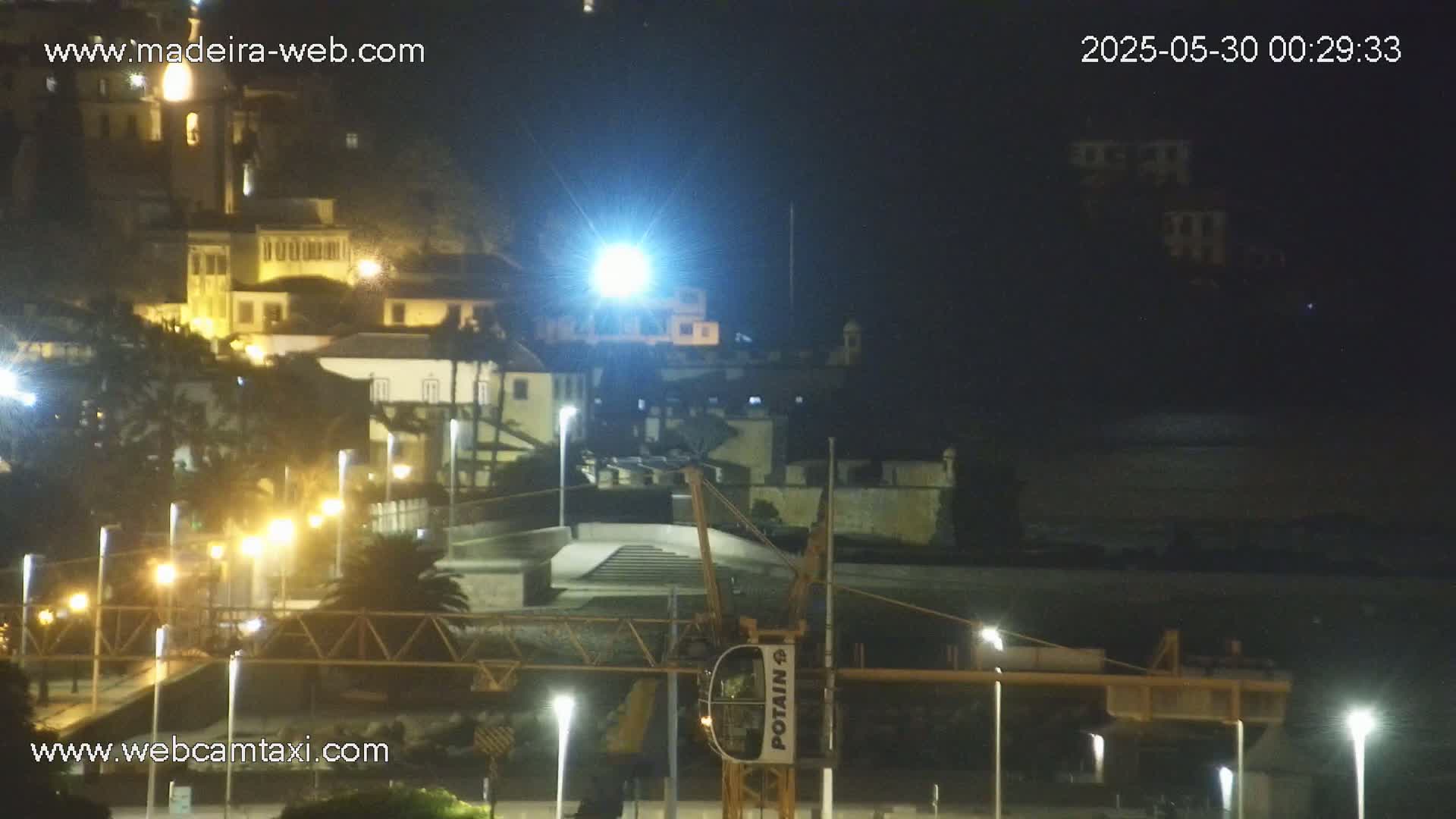 A nighttime view of a coastal town with illuminated buildings, streetlights, and a construction crane, under a dark sky.