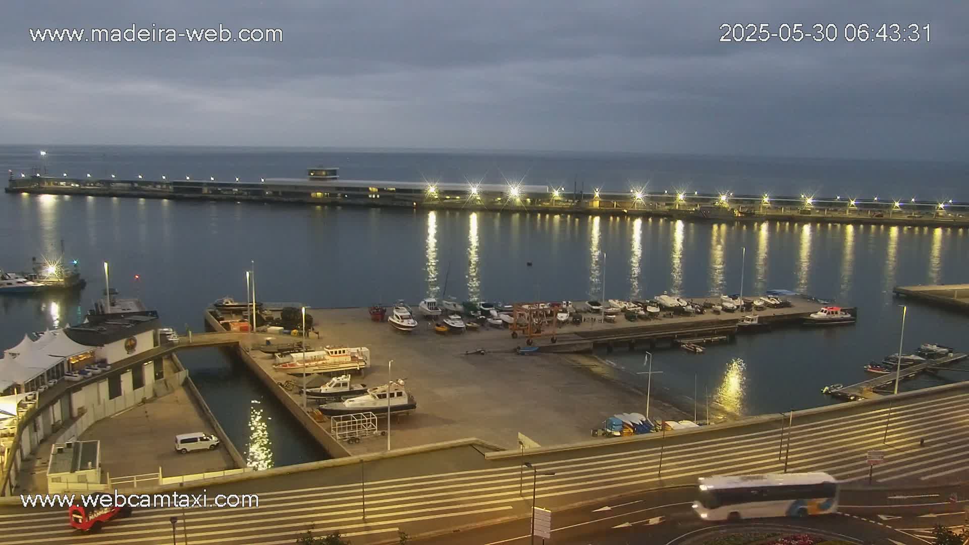 A harbor at twilight, with numerous boats docked, is illuminated by lights reflecting on the calm water under an overcast sky.