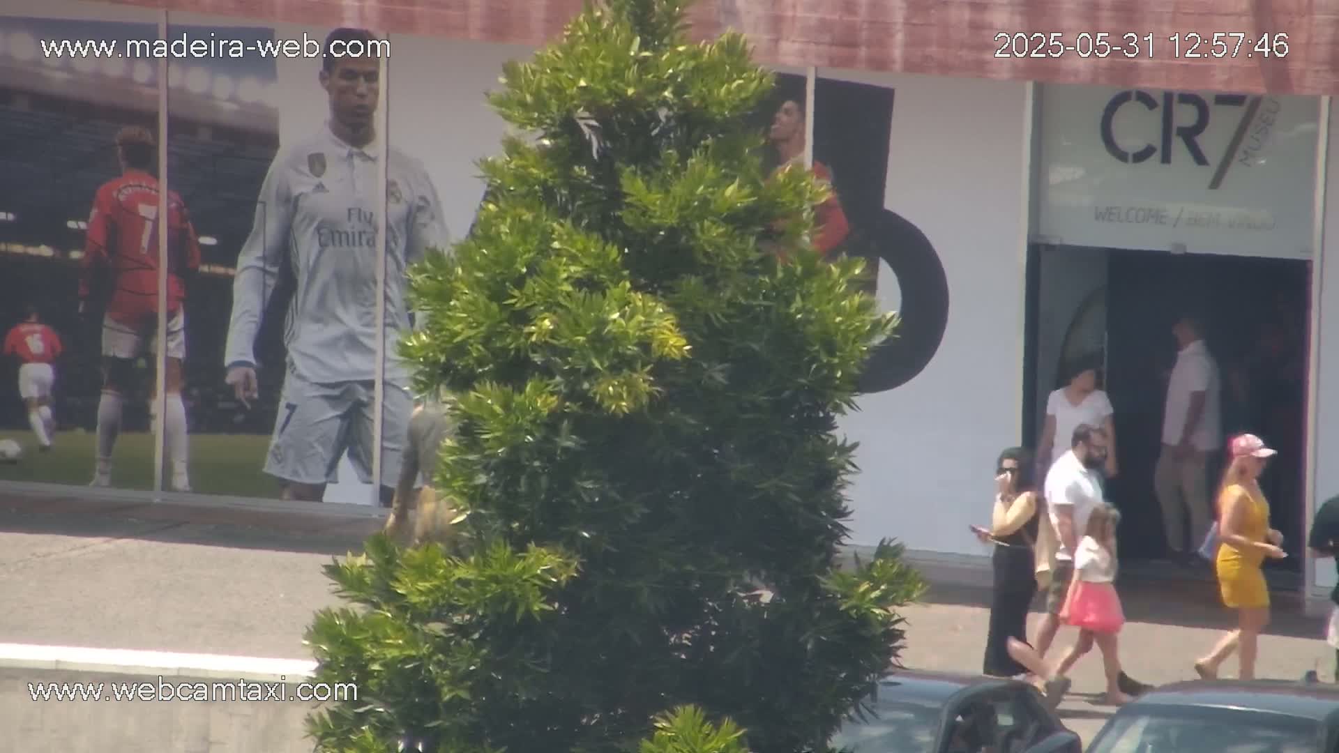A sunny day shows several people near a building with large images of Cristiano Ronaldo and other soccer players, and a green bush is in the foreground.