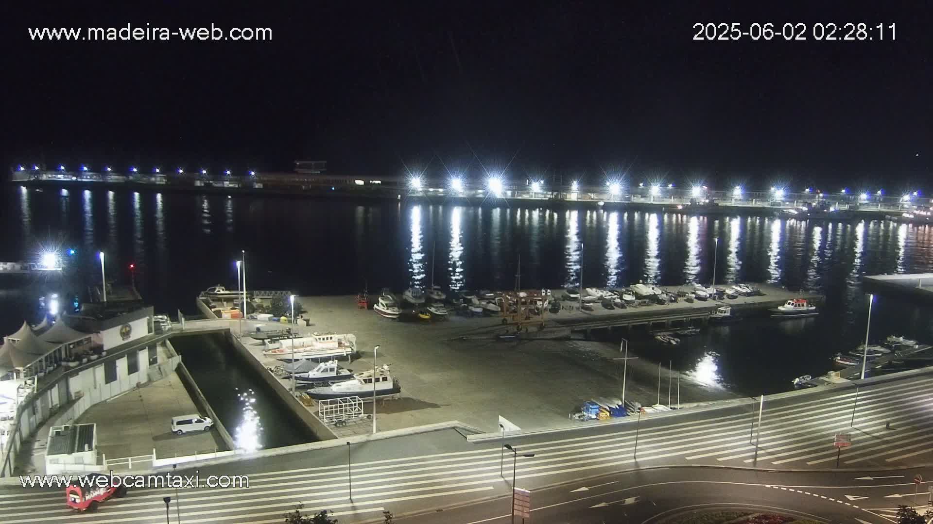A nighttime view of a harbor filled with boats, illuminated by bright lights reflecting on the calm water.