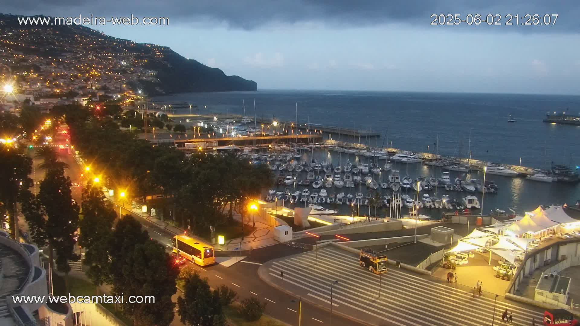 A nighttime view of a harbor filled with boats, next to a city illuminated with streetlights under a partly cloudy sky.