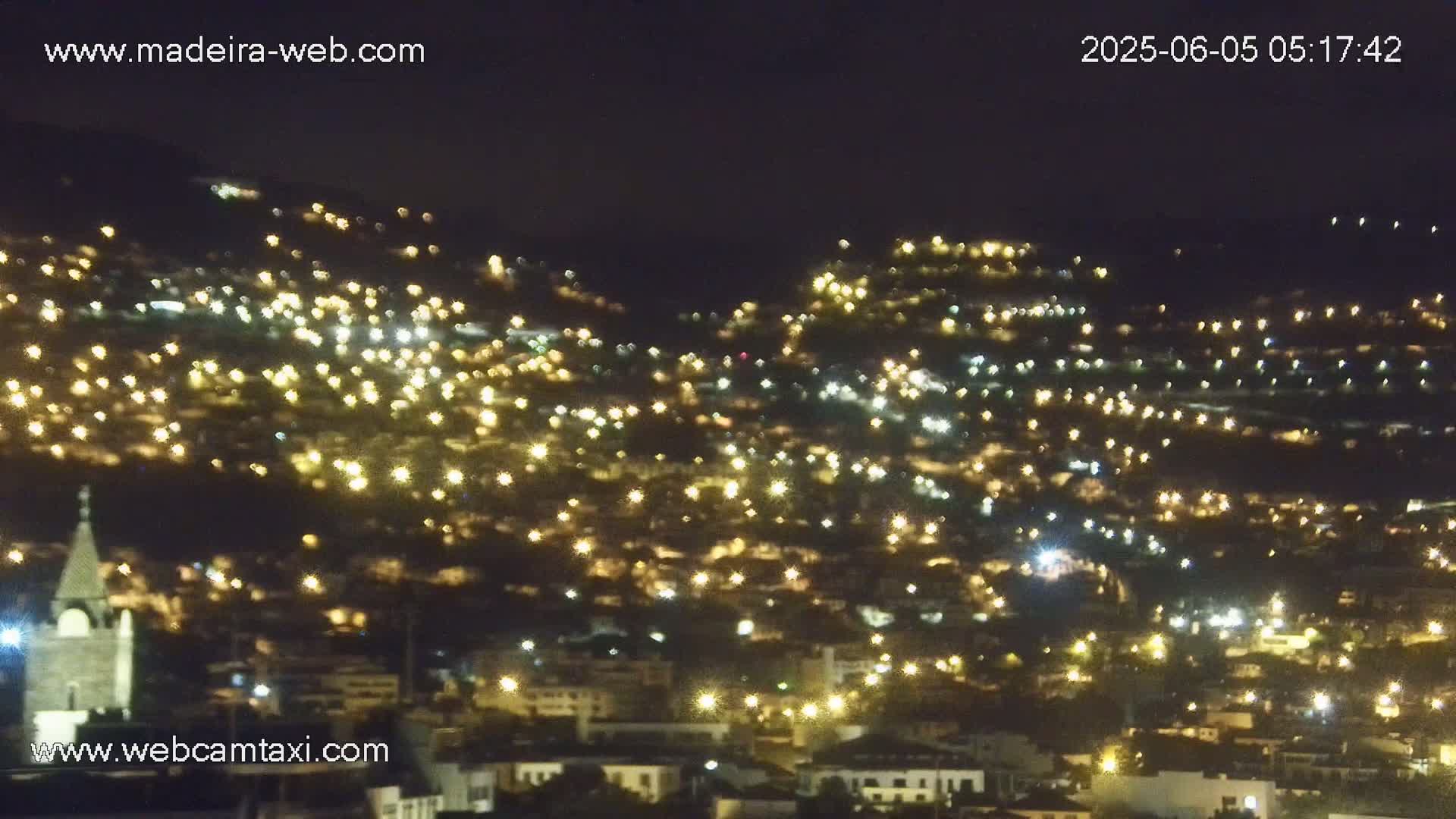 A hillside town at night is illuminated by numerous lights, appearing as a sparkling blanket across the landscape.