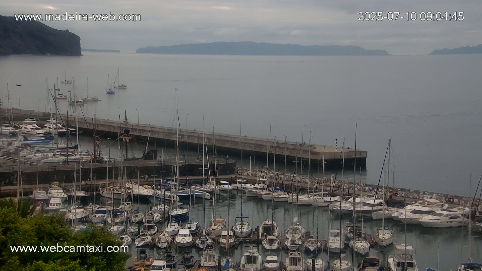 A marina filled with numerous sailboats and motorboats is situated next to a calm body of water under an overcast sky, with a distant cliff visible in the background.