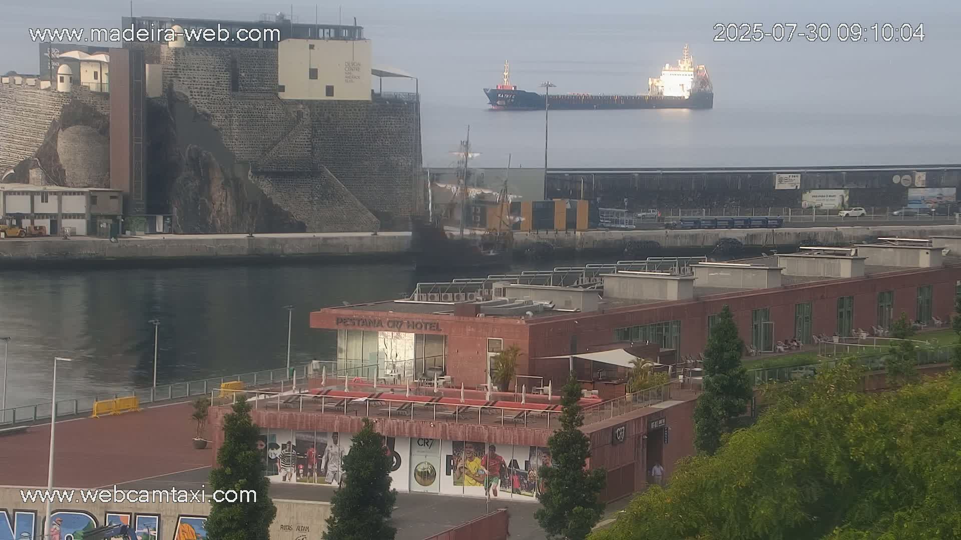 A harbor scene on a hazy day shows a large cargo ship in the distance, a hotel with a rooftop patio, and a stone embankment.
