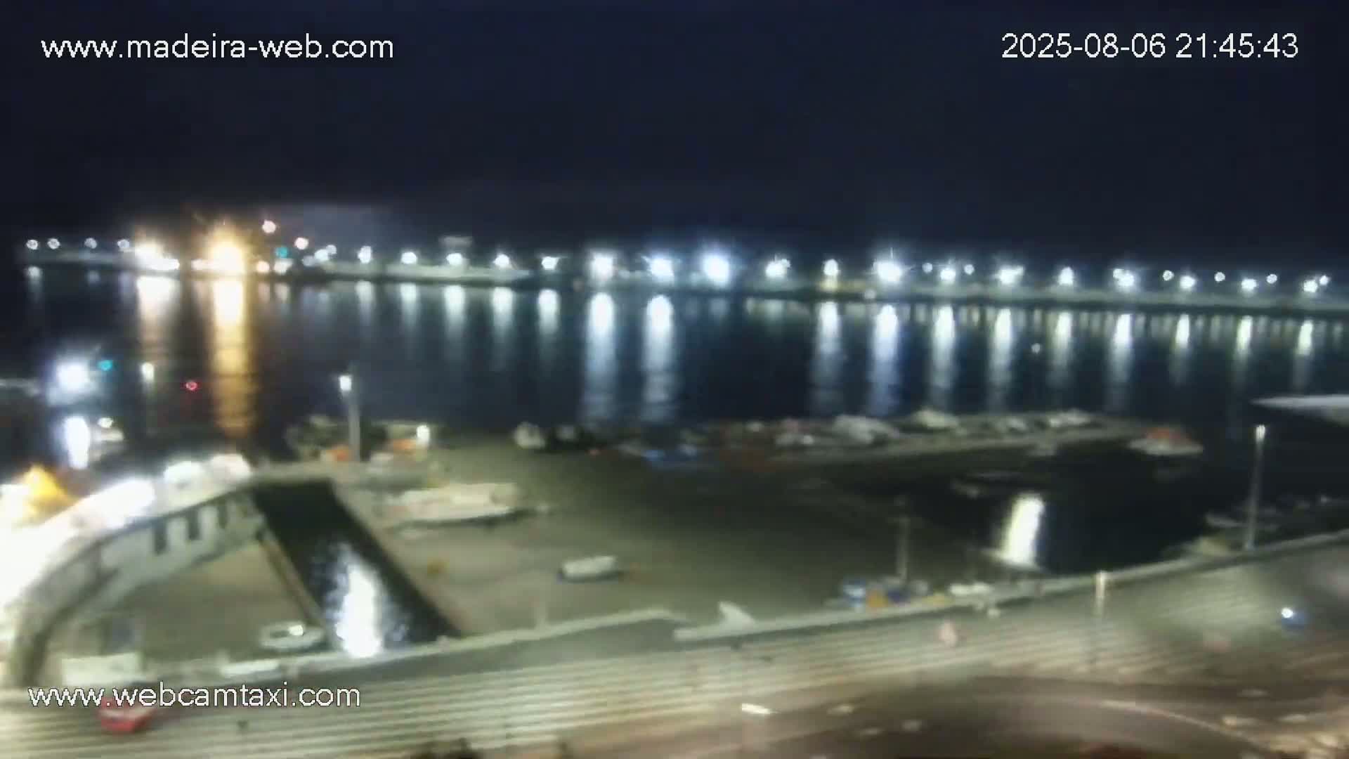 A nighttime view of a harbor, illuminated by lights reflecting on calm water, shows boats docked and a walkway leading down to the water.