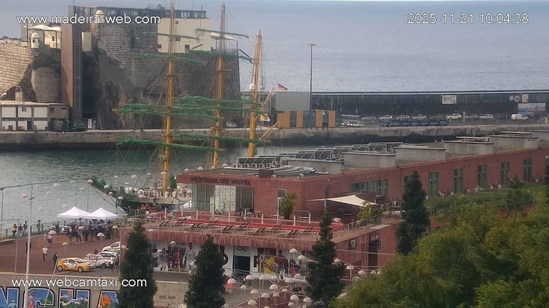 An overcast day at a bustling port features a large sailing ship docked near a lively promenade where people gather around white tents and several distinctive cars, with modern buildings and a rocky cliff face in the background and a calm sea stretching to the horizon.