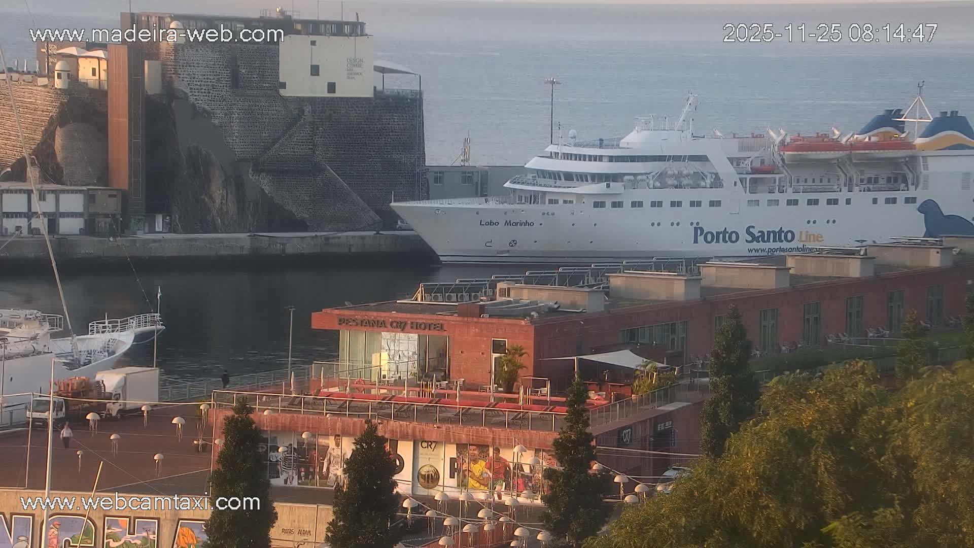 A large white "Porto Santo Line" ferry and another vessel named "Lobo Marinho" are docked in a calm harbor, flanked by waterfront buildings and rocky cliffs under clear, sunny morning skies.