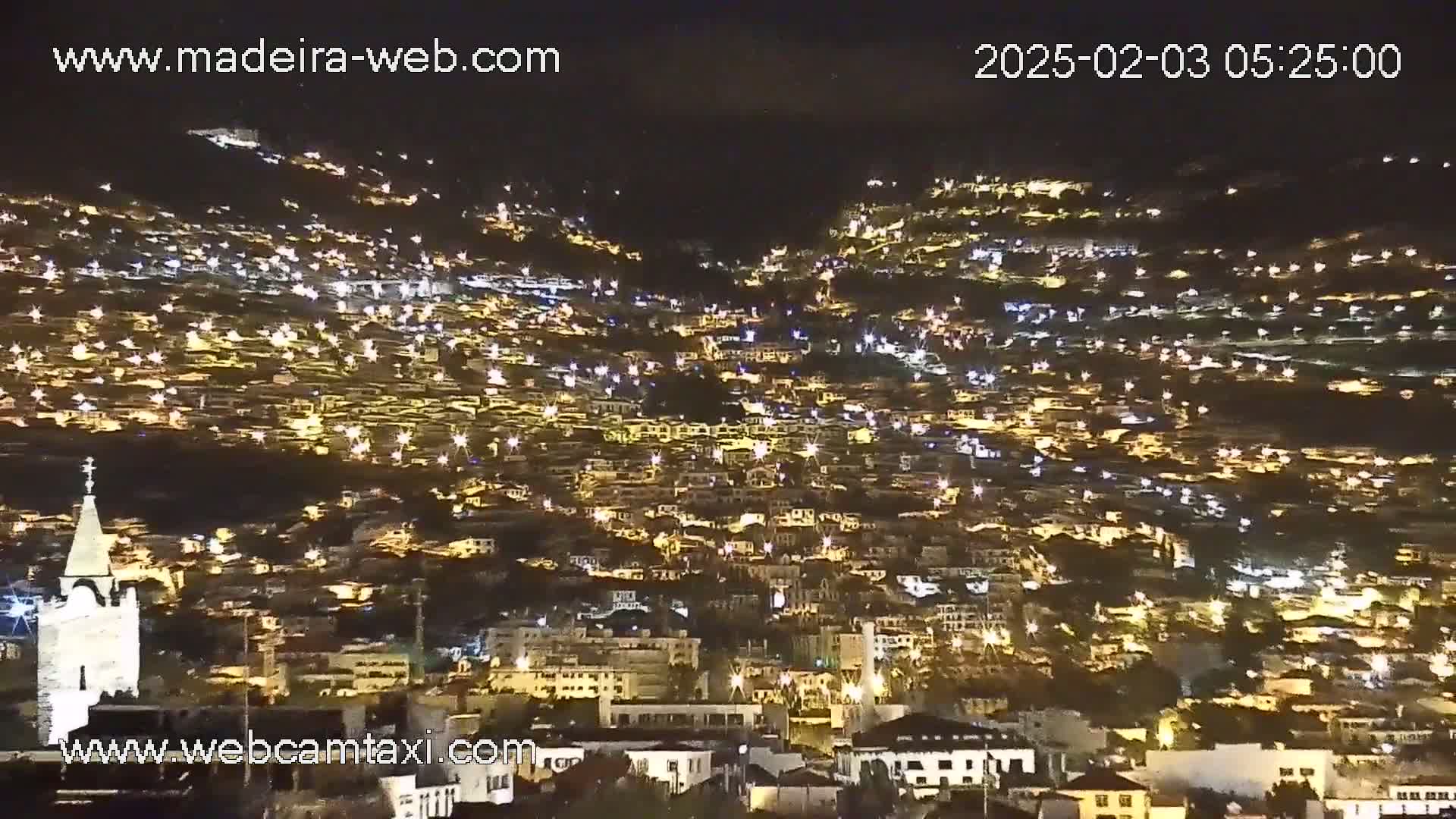 A nighttime, illuminated hillside town is seen from a distance under a clear, dark sky.