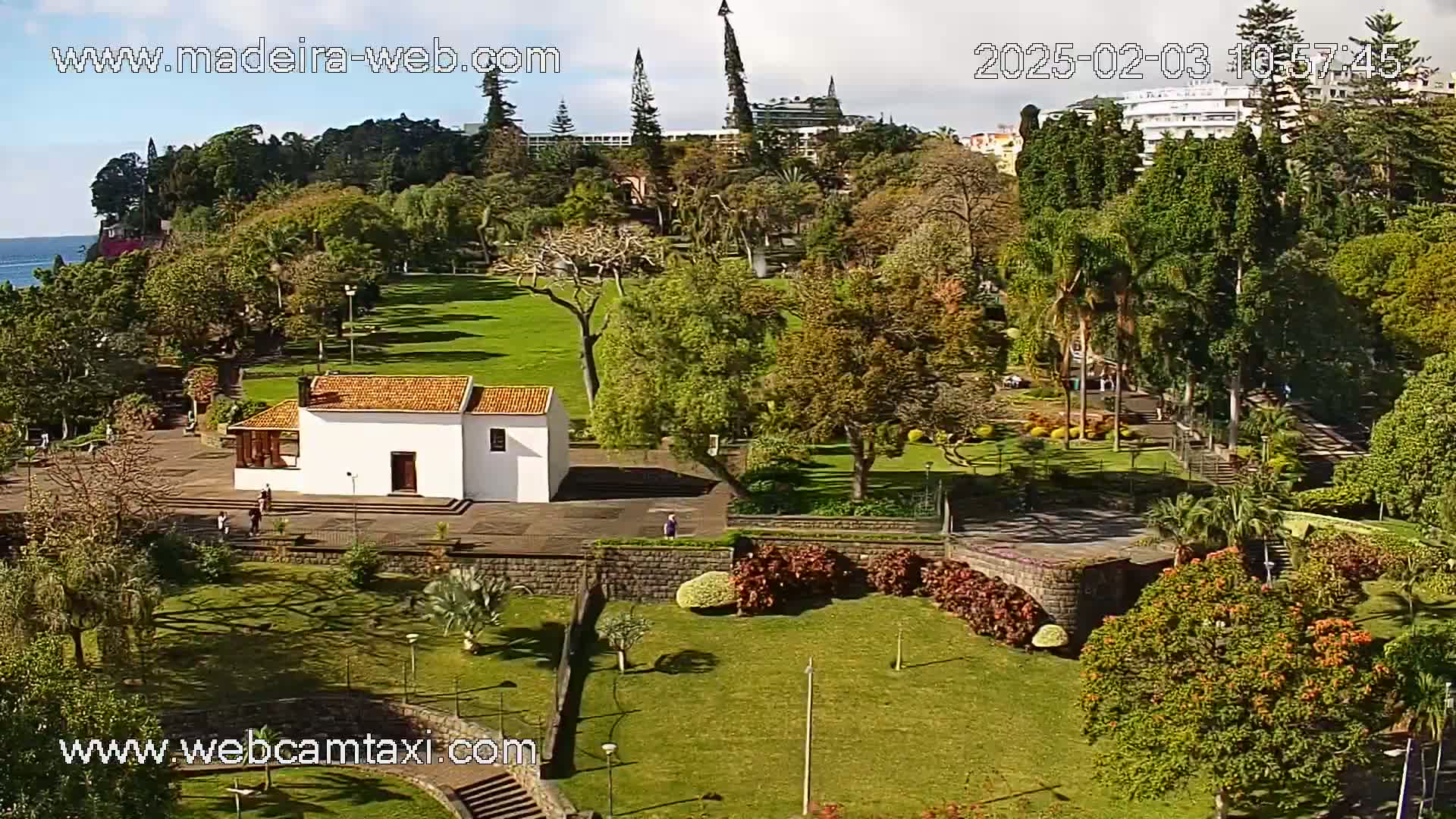 A high-angle view of a lush, sunny park with a small white building, surrounded by green lawns, flowering bushes, and various trees, with a partial view of buildings and the ocean in the distance.