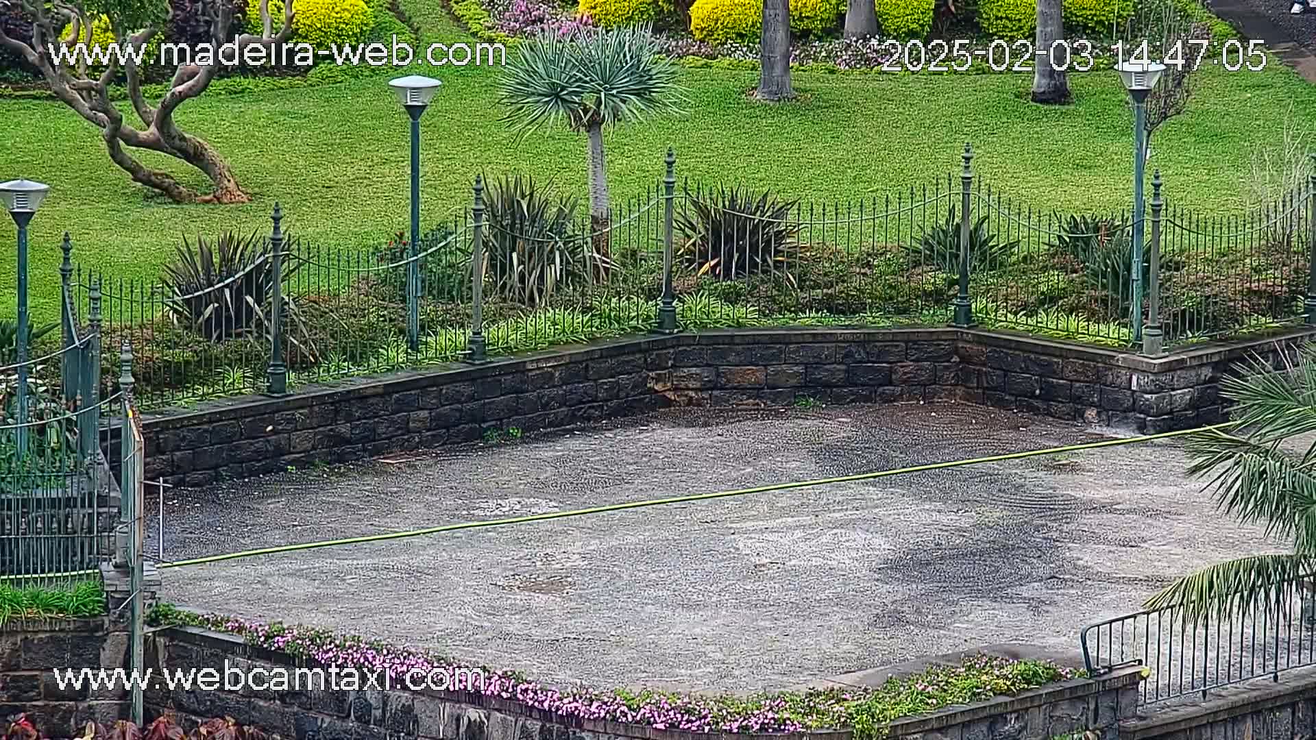A wet, paved area is surrounded by a low stone wall and a wrought-iron fence, with lush green lawns and various plants beyond.